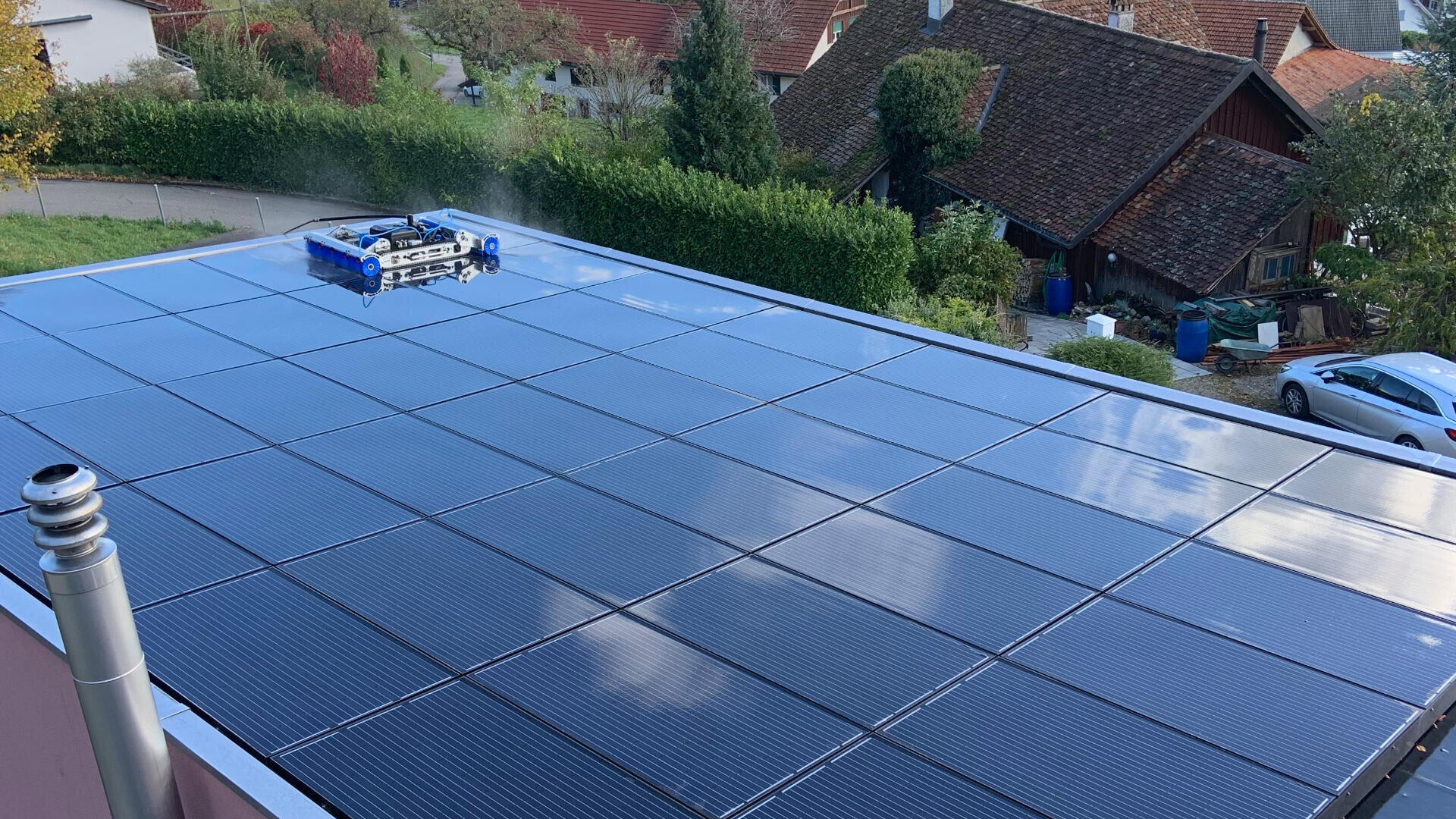 Ein mit Solarmodulen bedecktes Dach wird von einem kleinen Robotergerät gereinigt. Im Hintergrund sind Häuser, Bäume und Autos zu sehen.