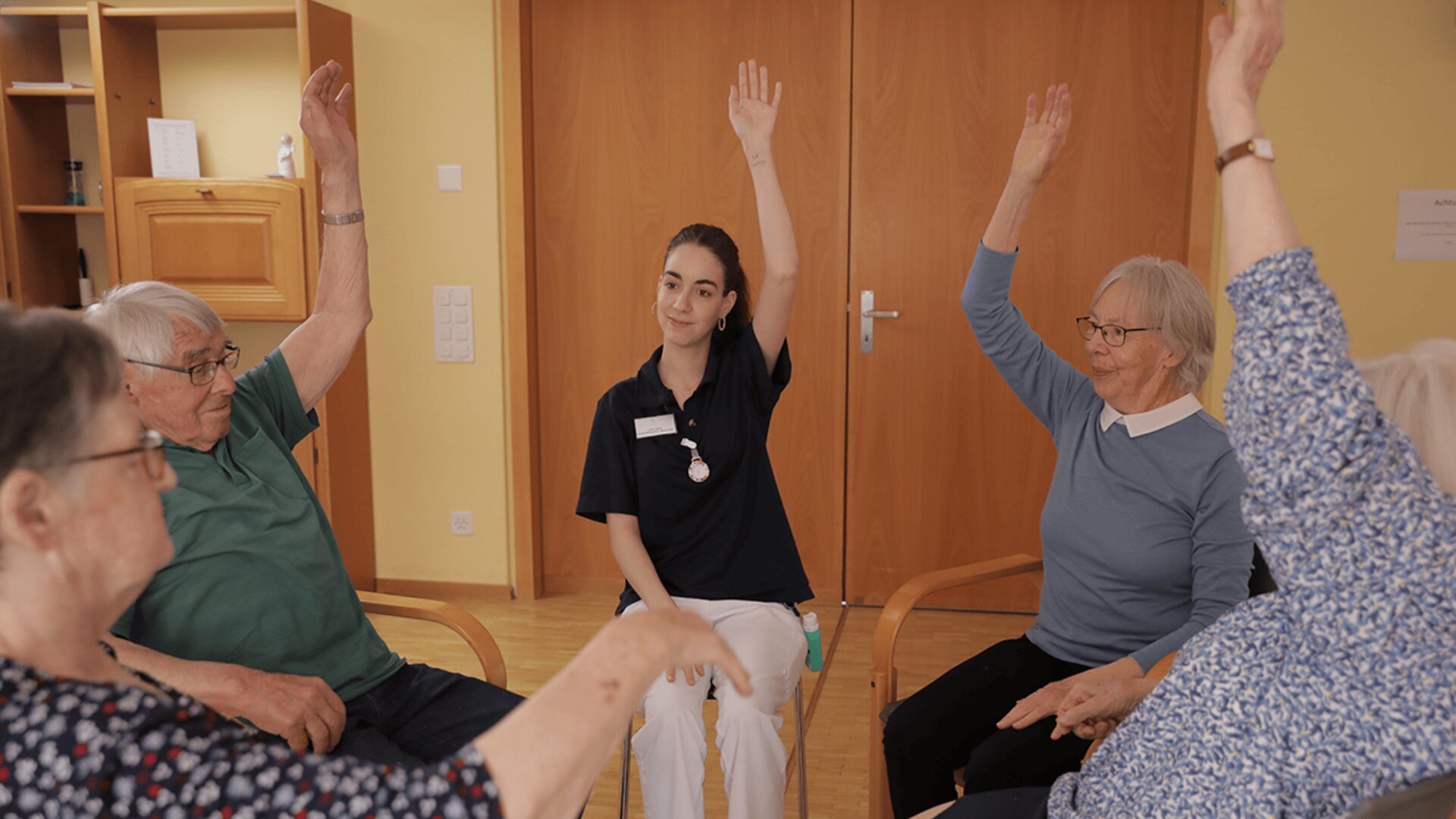 Eine Betreuerin und vier ältere Menschen sitzen im Kreis und heben während einer Gruppenaktivität in einem hell erleuchteten Raum die Hände. Die Atmosphäre wirkt freundlich und einnehmend.