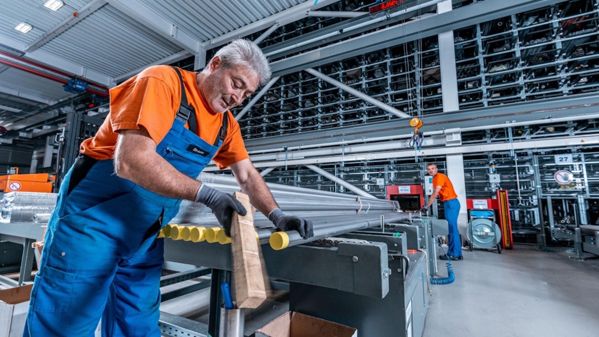 Ein älterer Mann in orange-blauer Arbeitskleidung hantiert in einer modernen Fabrik mit Metallrohren, während im Hintergrund ein anderer Arbeiter, umgeben von Industrieanlagen, eine Maschine bedient.