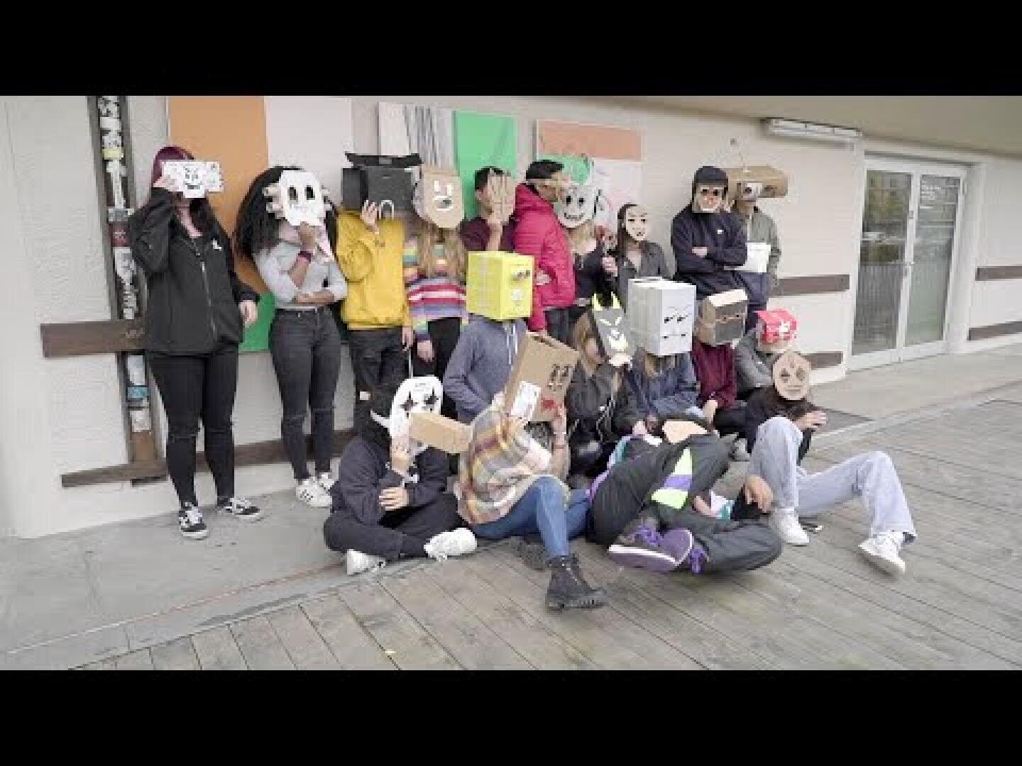 Eine Gruppe von Menschen posiert im Freien, jeder trägt eine große, handgefertigte Maske oder Kiste auf dem Kopf, die mit verschiedenen Farben und Mustern verziert ist. Einige stehen, andere sitzen oder liegen auf einem Holzdeck.