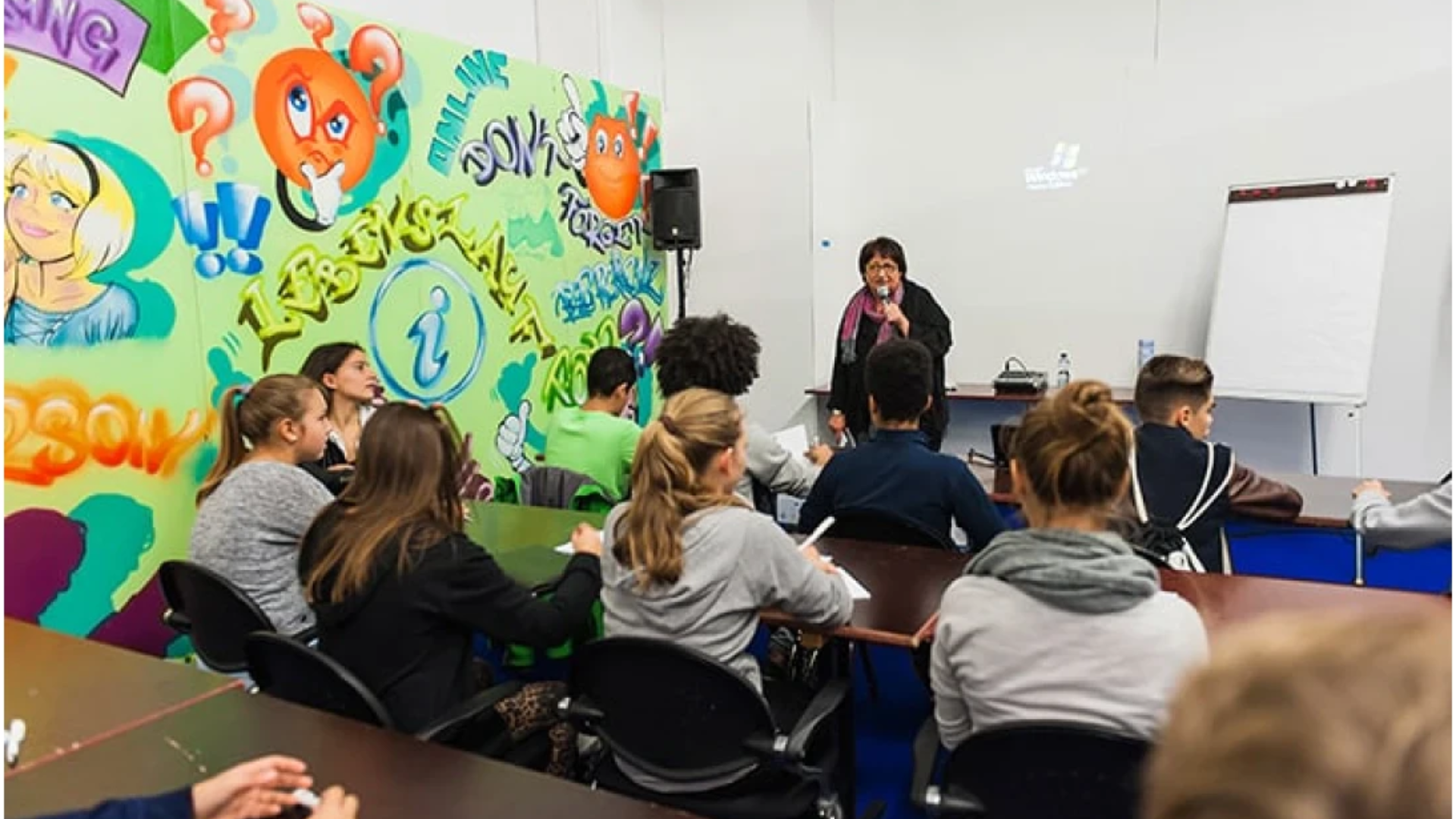 Eine Lehrerin steht vor einem Klassenzimmer und spricht mit ihren Schülern an den Tischen. Die Wand hinter ihr ist mit bunten Graffiti und Fragezeichen verziert. Ein Whiteboard und ein Lautsprecher sind ebenfalls zu sehen.