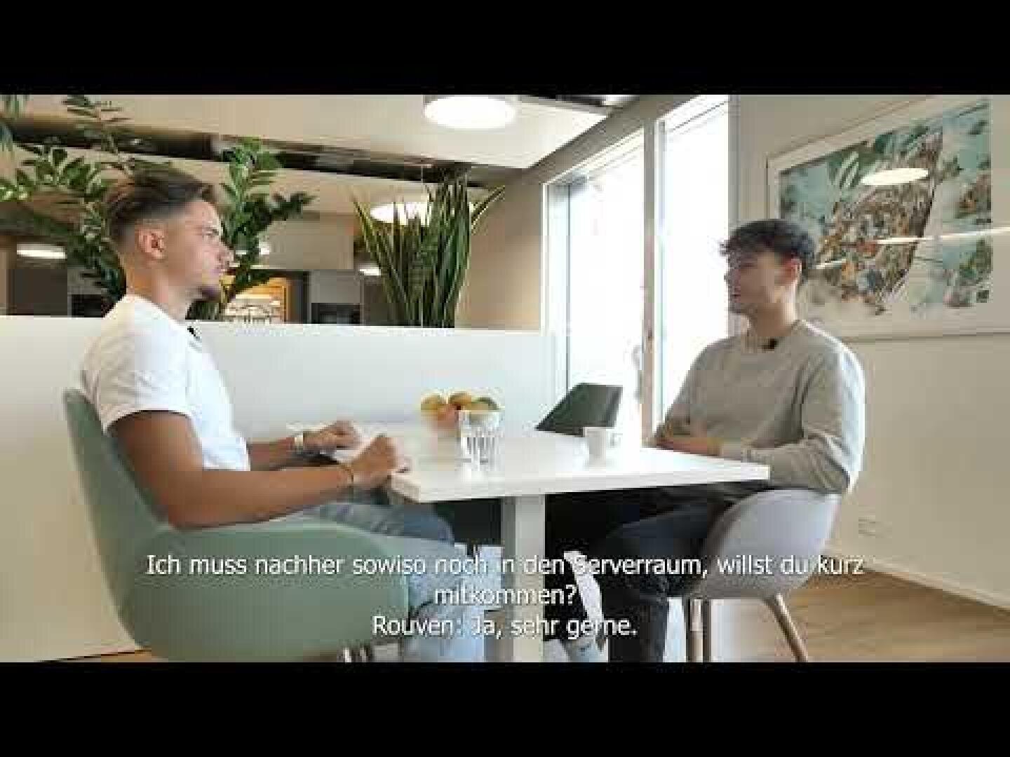 Zwei Personen sitzen sich an einem weißen Tisch gegenüber und unterhalten sich in einem modernen, gut beleuchteten Büro. Die deutschen Untertitel lauten: Ich muss nachher sowieso noch in den Serverraum, willst du kurz mitkommen? Rouven: Ja, sehr gerne.
