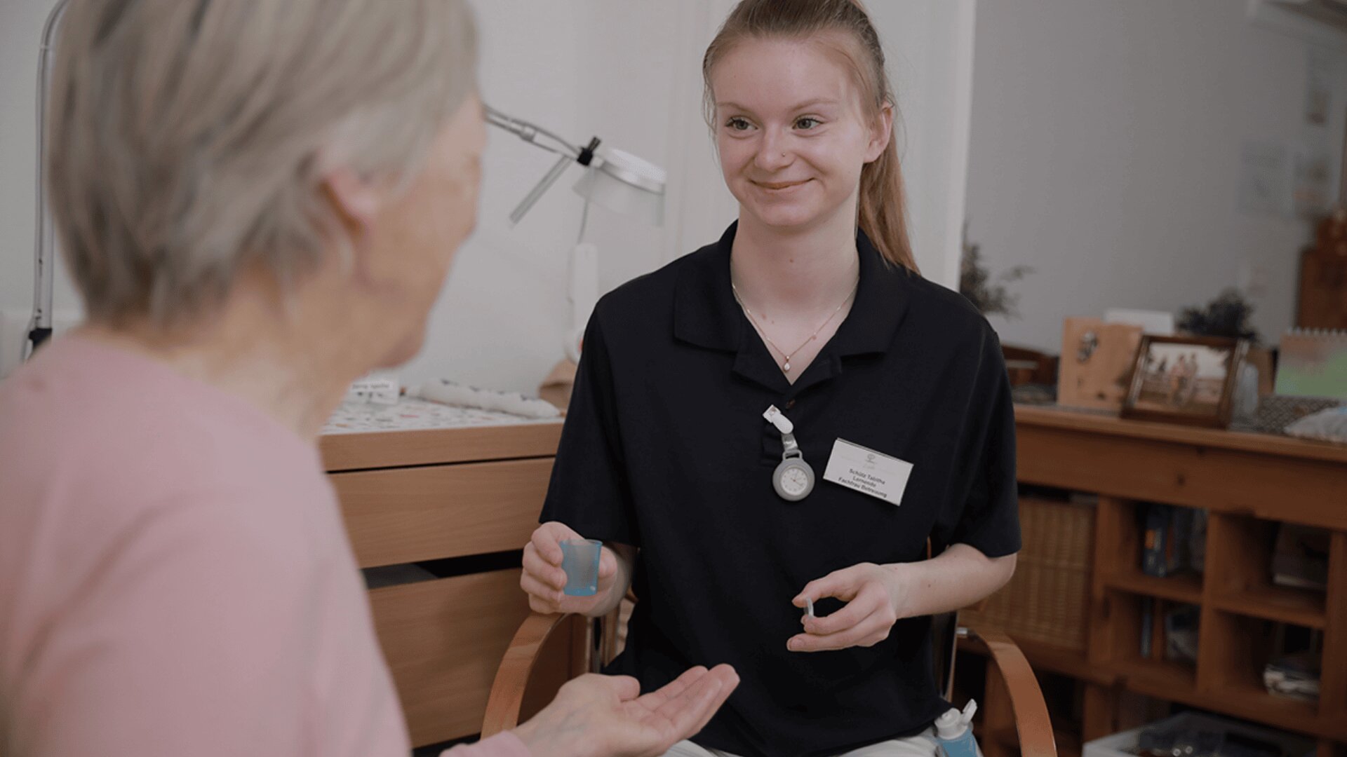 Eine junge Pflegerin mit marineblauem Hemd und Namensschild lächelt, während sie einer älteren Frau mit grauem Haar, die in einem gemütlichen, gut beleuchteten Raum sitzt und ihre Hand ausstreckt, Medikamente reicht.