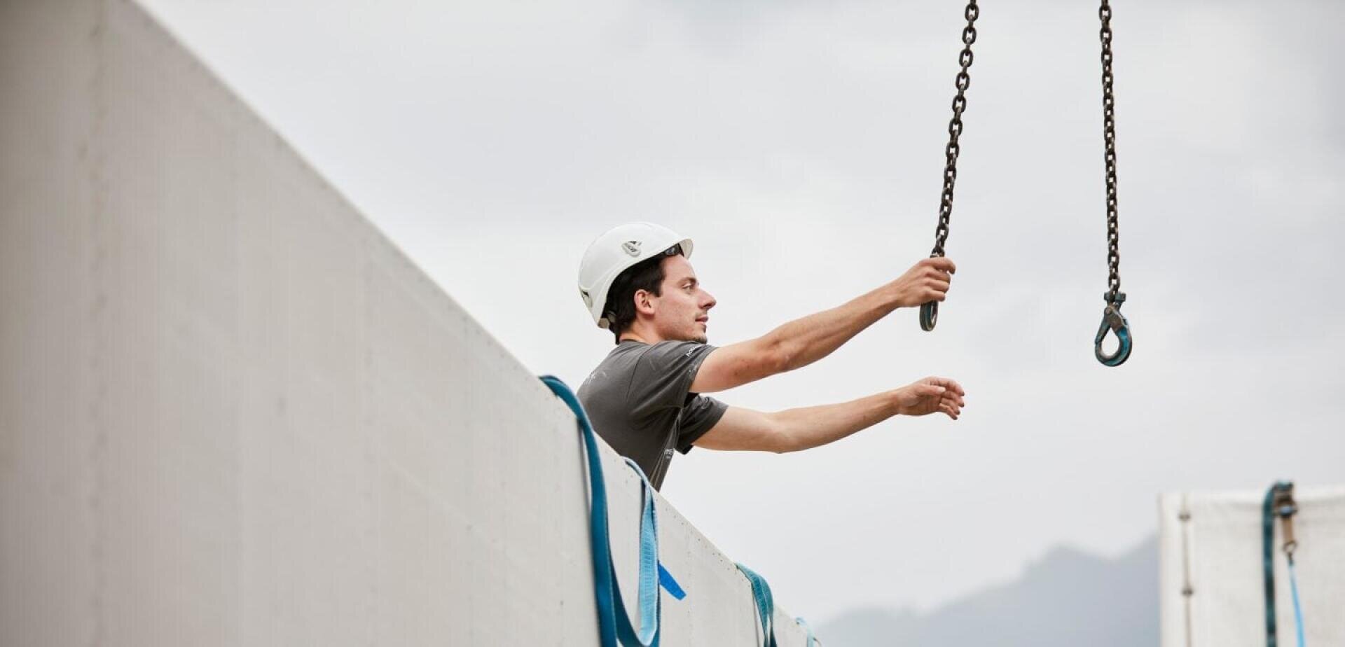 Ein Bauarbeiter mit Schutzhelm greift nach einer Kran-Kette, die über einer Betonmauer hängt.