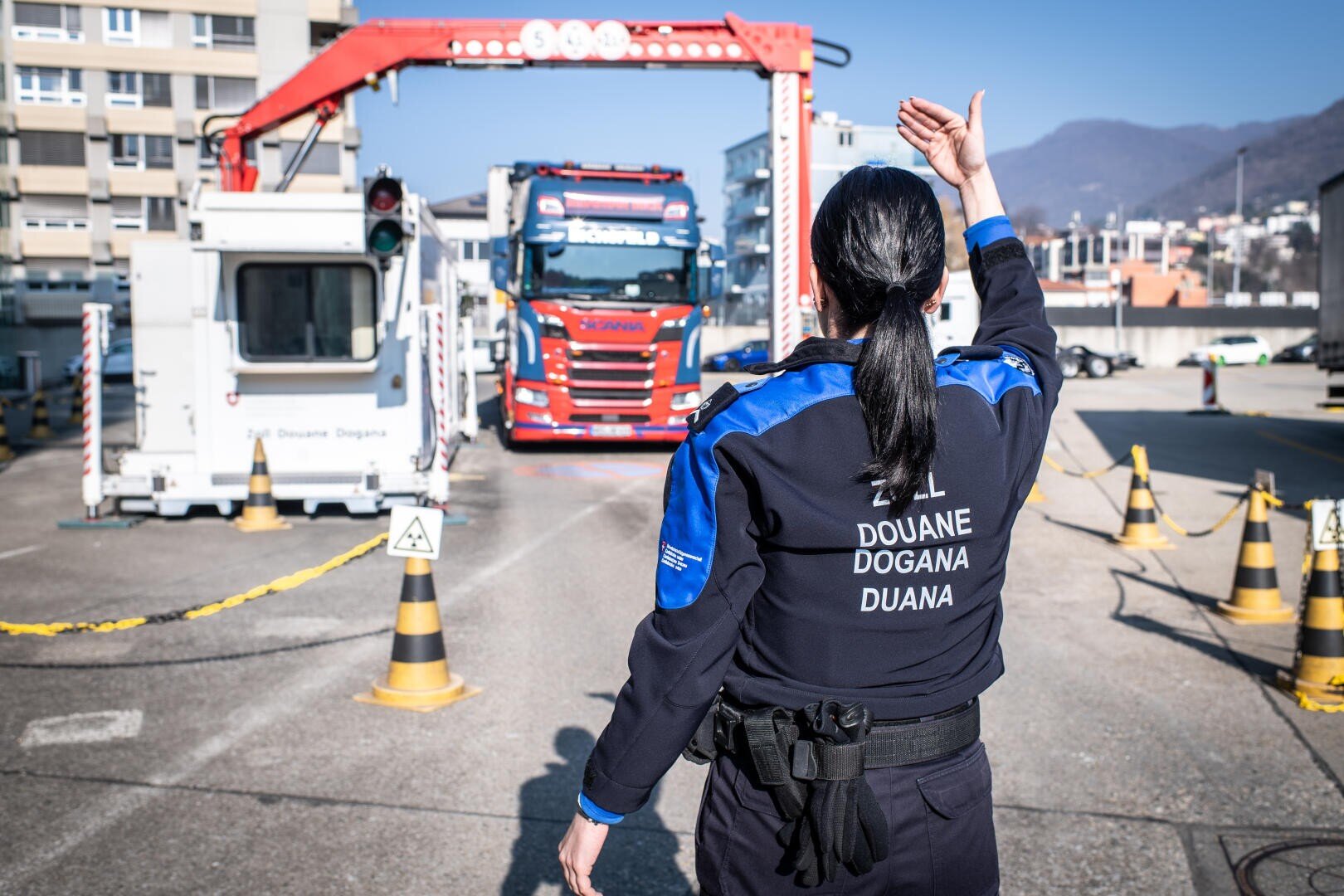 Eine Zollbeamtin steht mit dem Rücken zur Kamera und hebt ihre Hand, um einem roten Lastwagen zu signalisieren, an einem Grenzkontrollpunkt anzuhalten. Im Hintergrund sind Gebäude und Berge zu sehen.