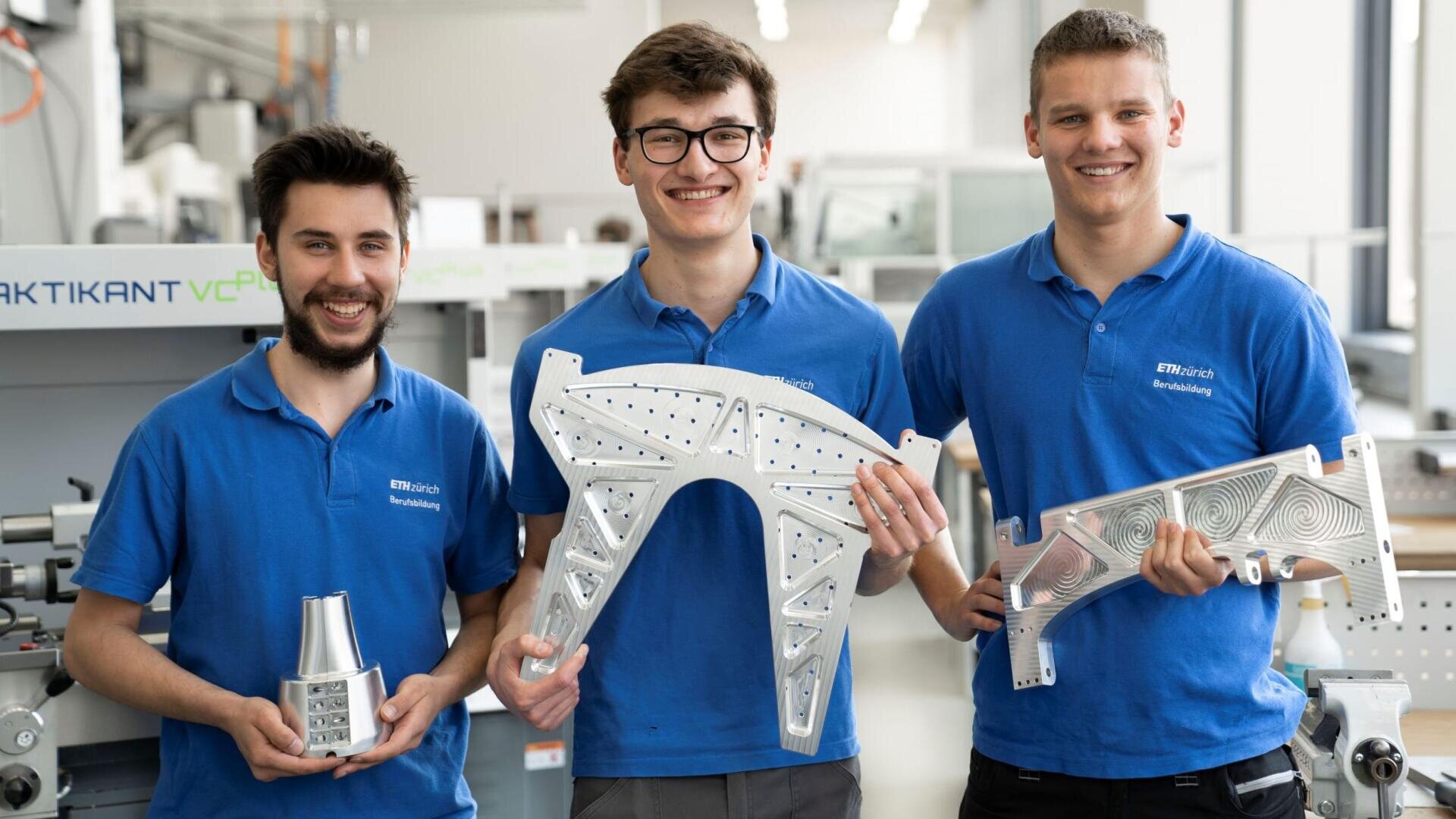 Drei junge Männer in blauen Hemden stehen lächelnd in einer Werkstatt und halten große, metallische, bearbeitete Teile in den Händen. Im hellen Hintergrund sind Industrieanlagen und Arbeitsplätze zu sehen.