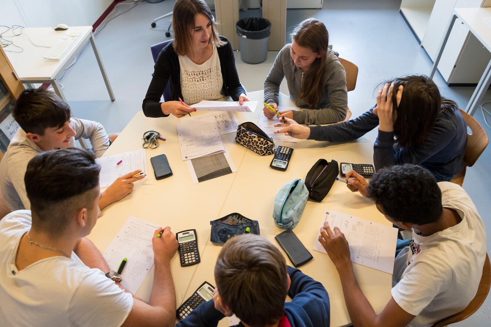 Sieben Schüler sitzen mit Papieren, Taschenrechnern und Federmäppchen um einen Tisch herum und arbeiten gemeinsam in einem Klassenzimmer. Sie scheinen gerade an einer Gruppendiskussion oder einer gemeinsamen Lernsitzung teilzunehmen.