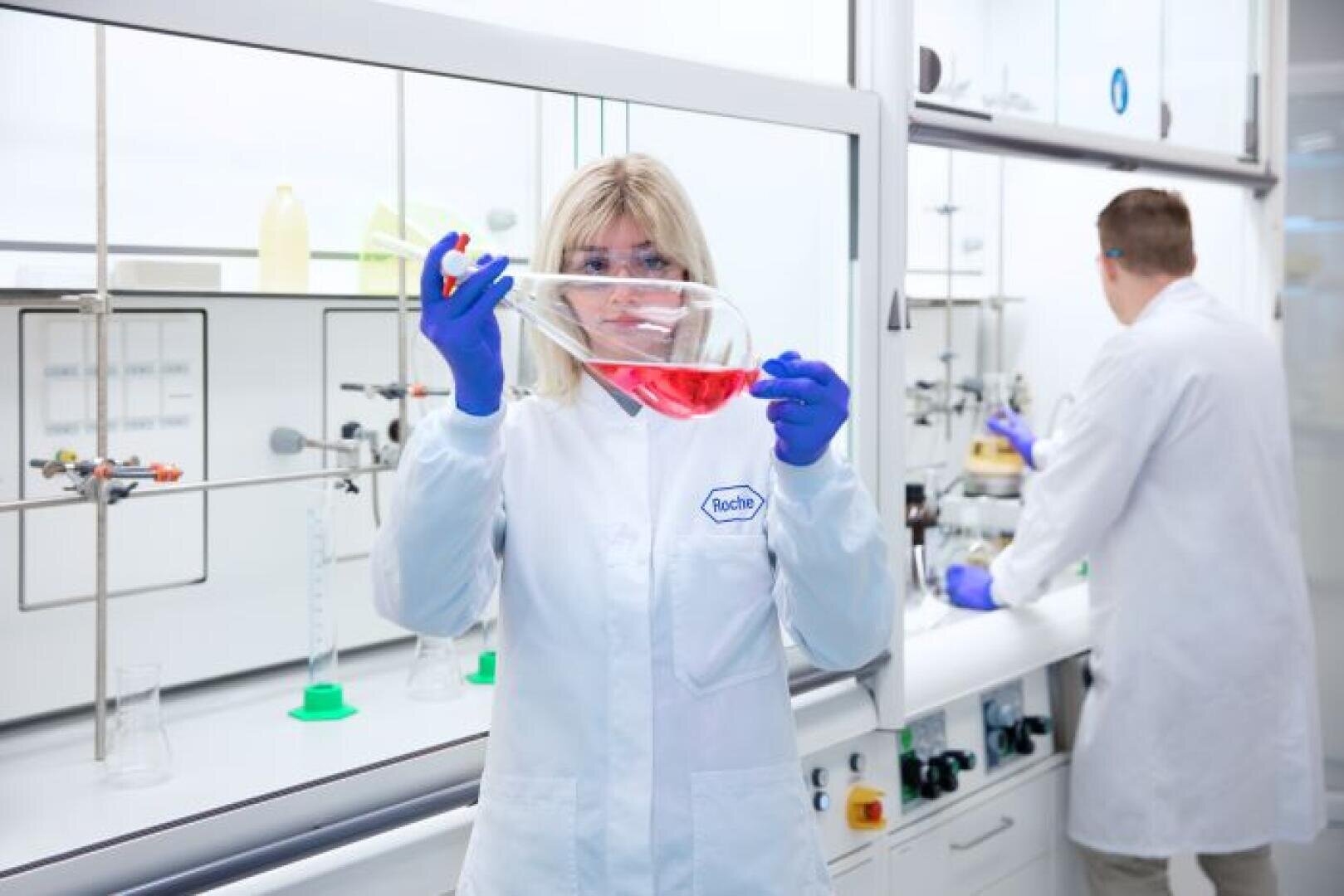 Eine Wissenschaftlerin in einem Laborkittel und mit blauen Handschuhen untersucht in einem modernen Labor einen großen Becher mit roter Flüssigkeit, während im Hintergrund ein männlicher Wissenschaftler arbeitet.