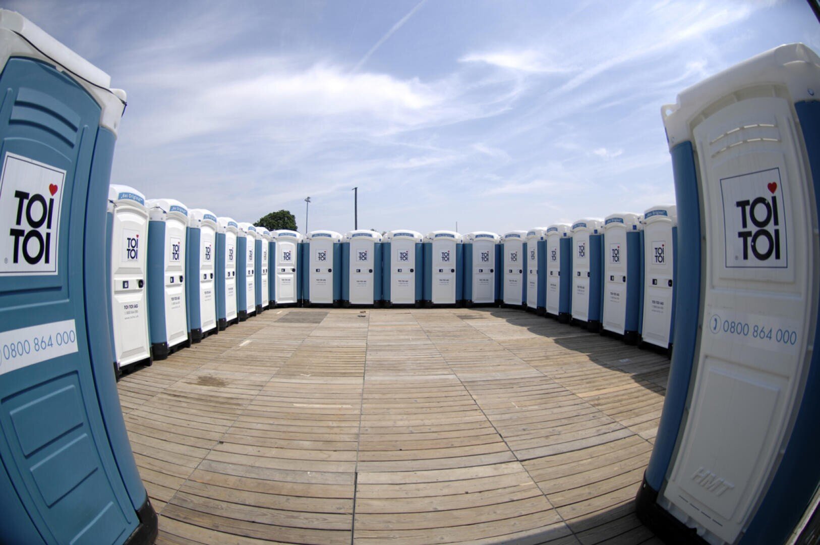 Unter einem teilweise bewölkten Himmel steht im Freien auf einer Holzplattform eine Reihe blau-weißer Mobiltoiletten.