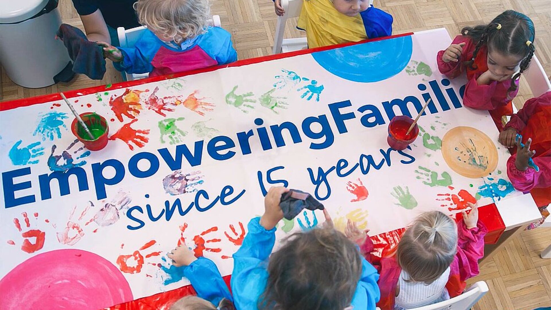 Kinder in farbenfrohen Schürzen machen Handabdrücke auf einem großen Banner mit der Aufschrift "Empowering Families since 15 years" (Familien stärken seit 15 Jahren) und benutzen dazu Farben und Pinsel an einem Tisch in einem hellen, freundlichen Raum.