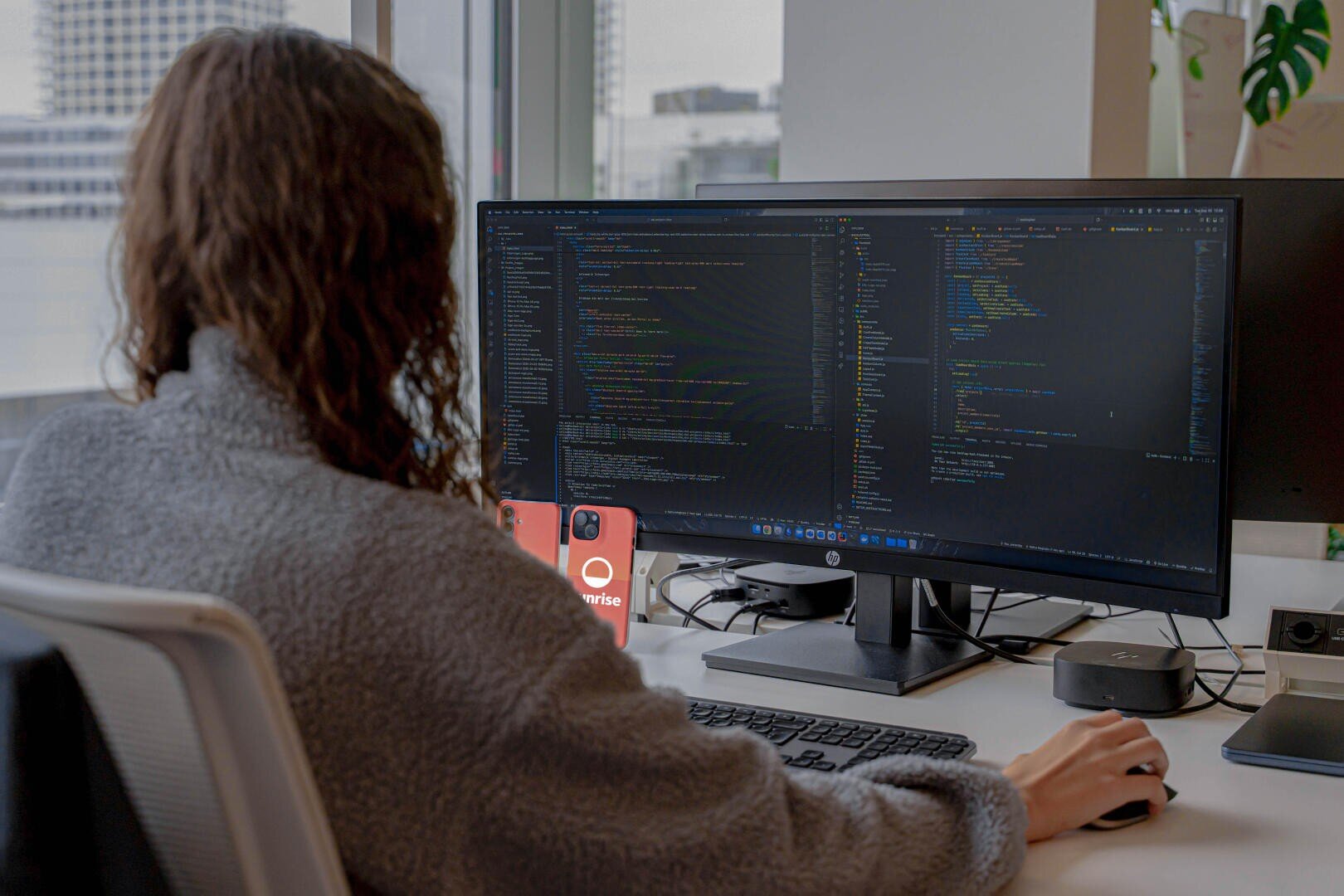 Eine Person mit langen lockigen Haaren sitzt an einem Schreibtisch und verwendet einen großen Monitor, auf dem Codes angezeigt werden. Auf dem Schreibtisch liegen zwei rote Smartphones, und der Arbeitsplatz befindet sich an einem Fenster mit Blick auf städtische Gebäude.