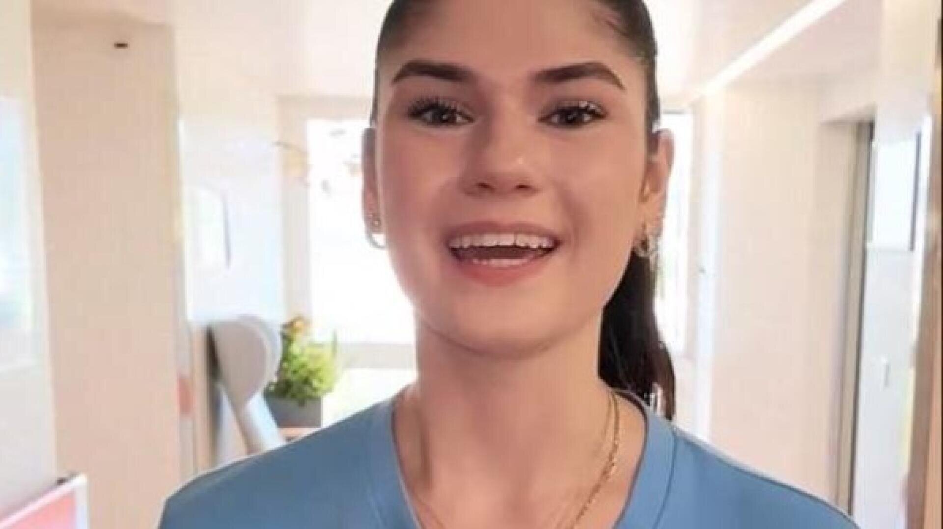 Eine junge Frau mit langen, dunklen Haaren, die zu einem Zopf zusammengebunden sind, und einem blauen Hemd lächelt herzlich, während sie in einem hell erleuchteten Raum steht.