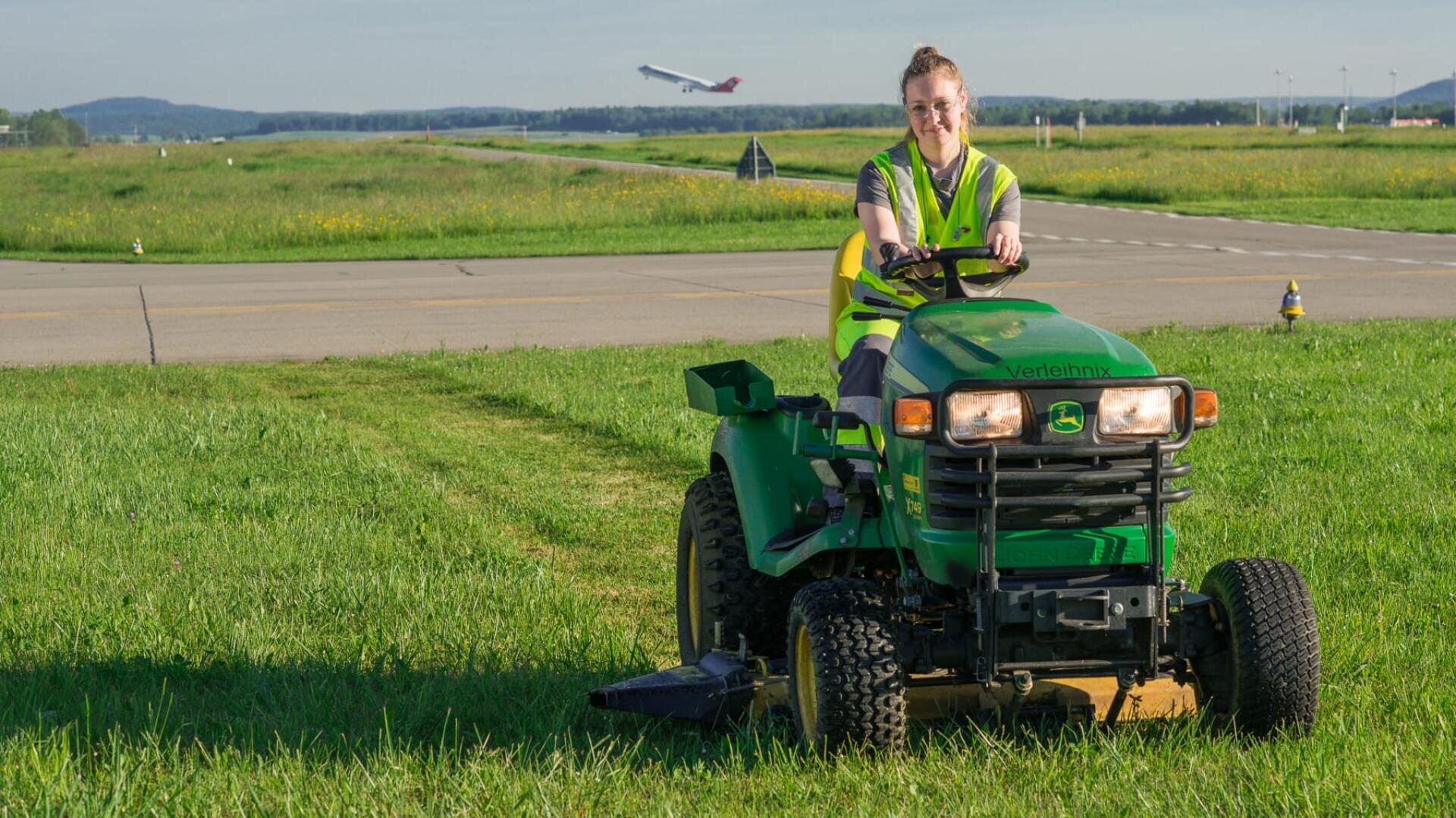 Eine Frau in einer Warnweste fährt einen grünen Rasenmäher auf einer Wiese in der Nähe einer Start- und Landebahn eines Flughafens, während im Hintergrund ein Flugzeug abhebt.