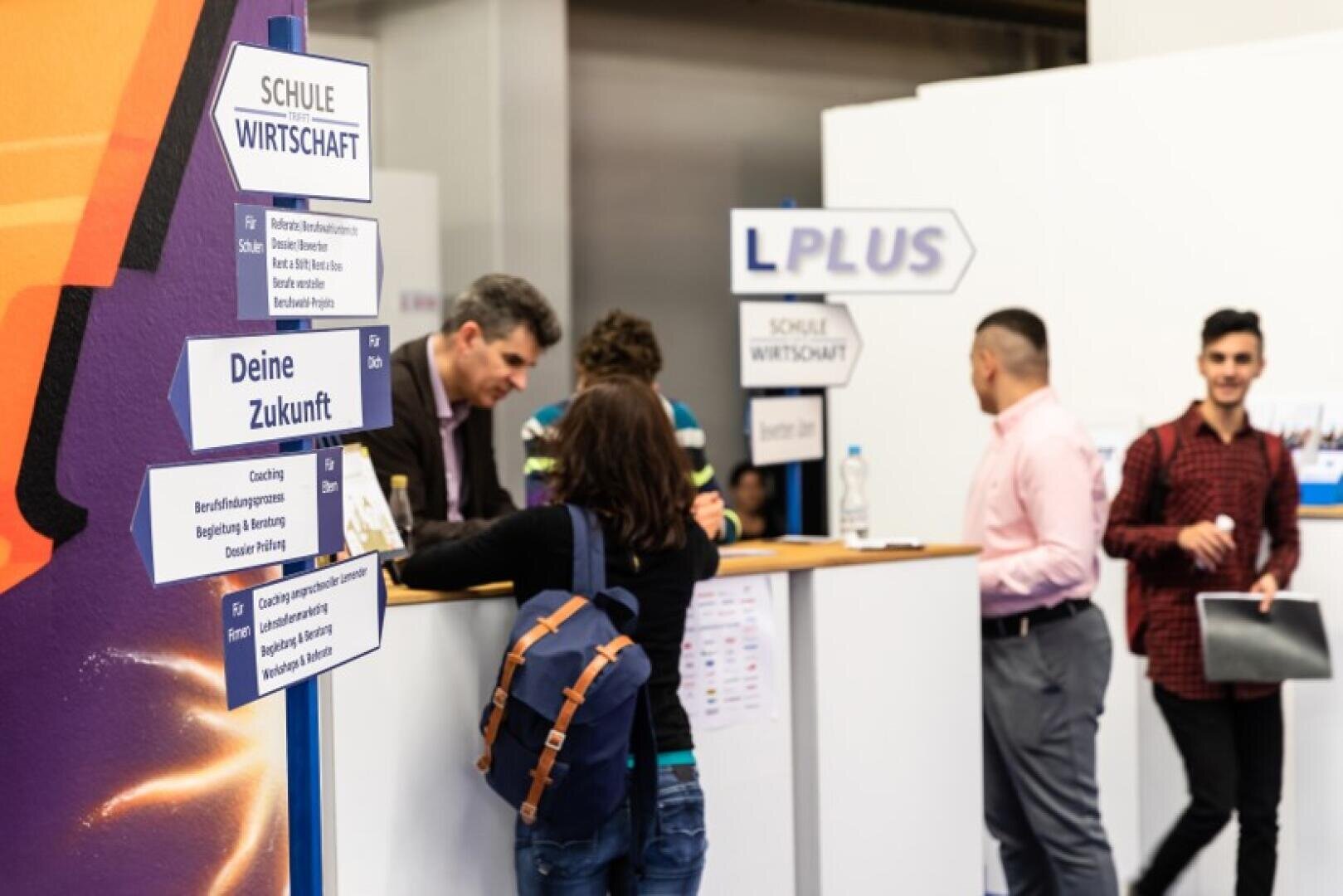 Menschen unterhalten sich an einem Informationsstand auf einer Karrieremesse. Auf den Schildern auf Deutsch steht Schule Wirtschaft und Deine Zukunft. Mehrere Personen stellen Fragen, während die Mitarbeiter Informationen geben.