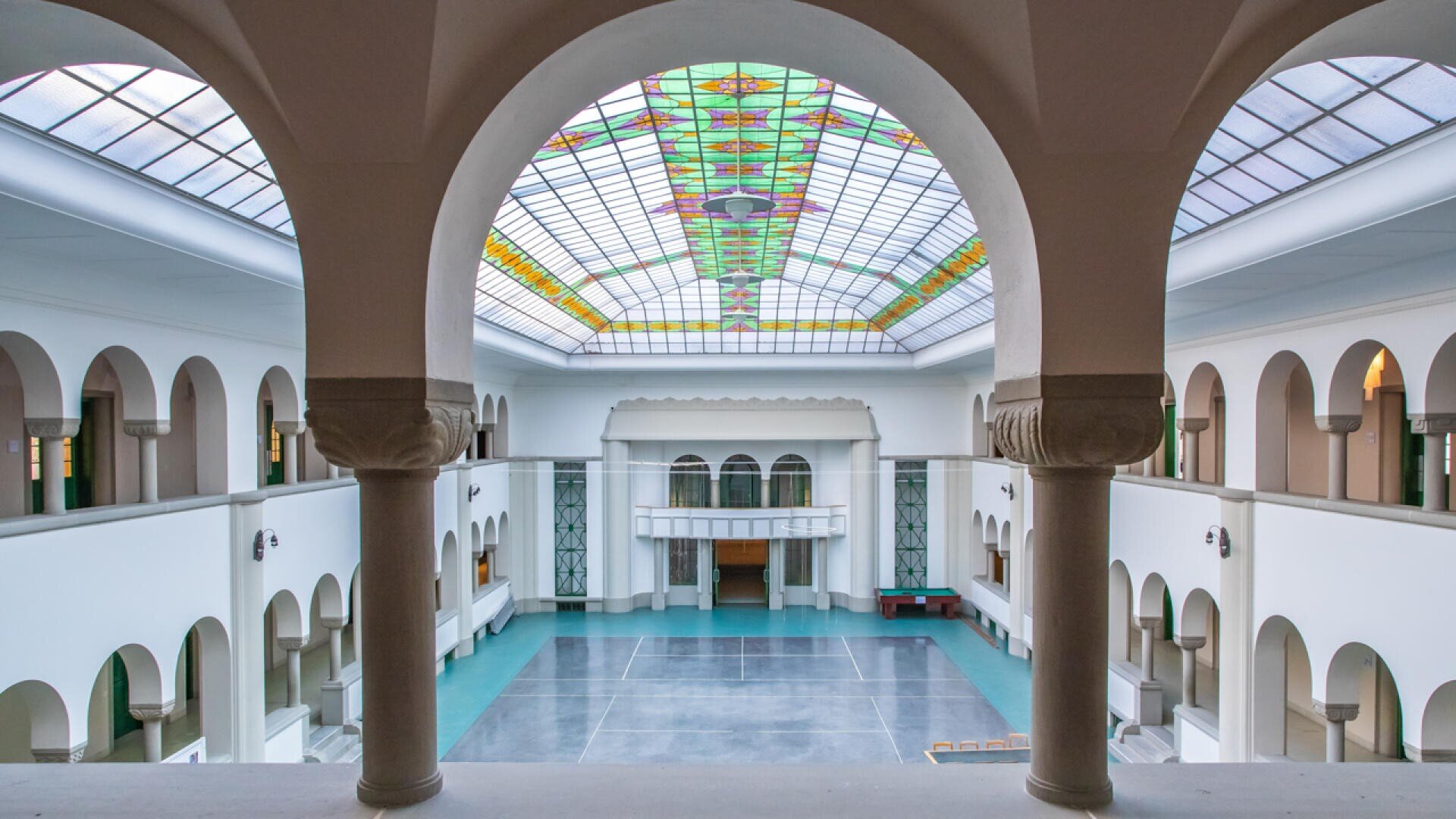 Geräumiger Innenhof mit gewölbten Säulen, einer gemusterten Glasdecke und einem zentralen offenen Bereich, der von weißen Wänden und grünen Akzenten umgeben ist. Natürliches Licht strömt durch das bunte Glas darüber.