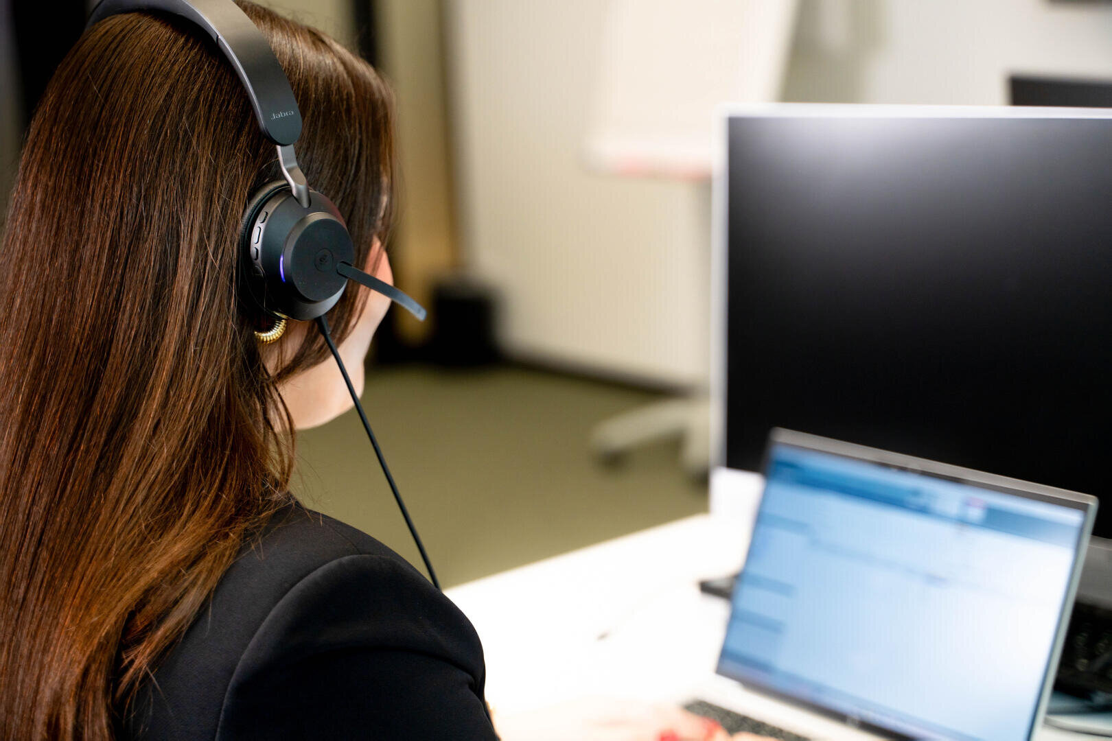 Eine Person mit langen braunen Haaren, die ein Headset trägt, sitzt an einem Schreibtisch, blickt auf Computerbildschirme und arbeitet an einem Laptop; möglicherweise führt sie gerade ein Videogespräch oder ist im Kundensupport in einer Büroumgebung tätig.