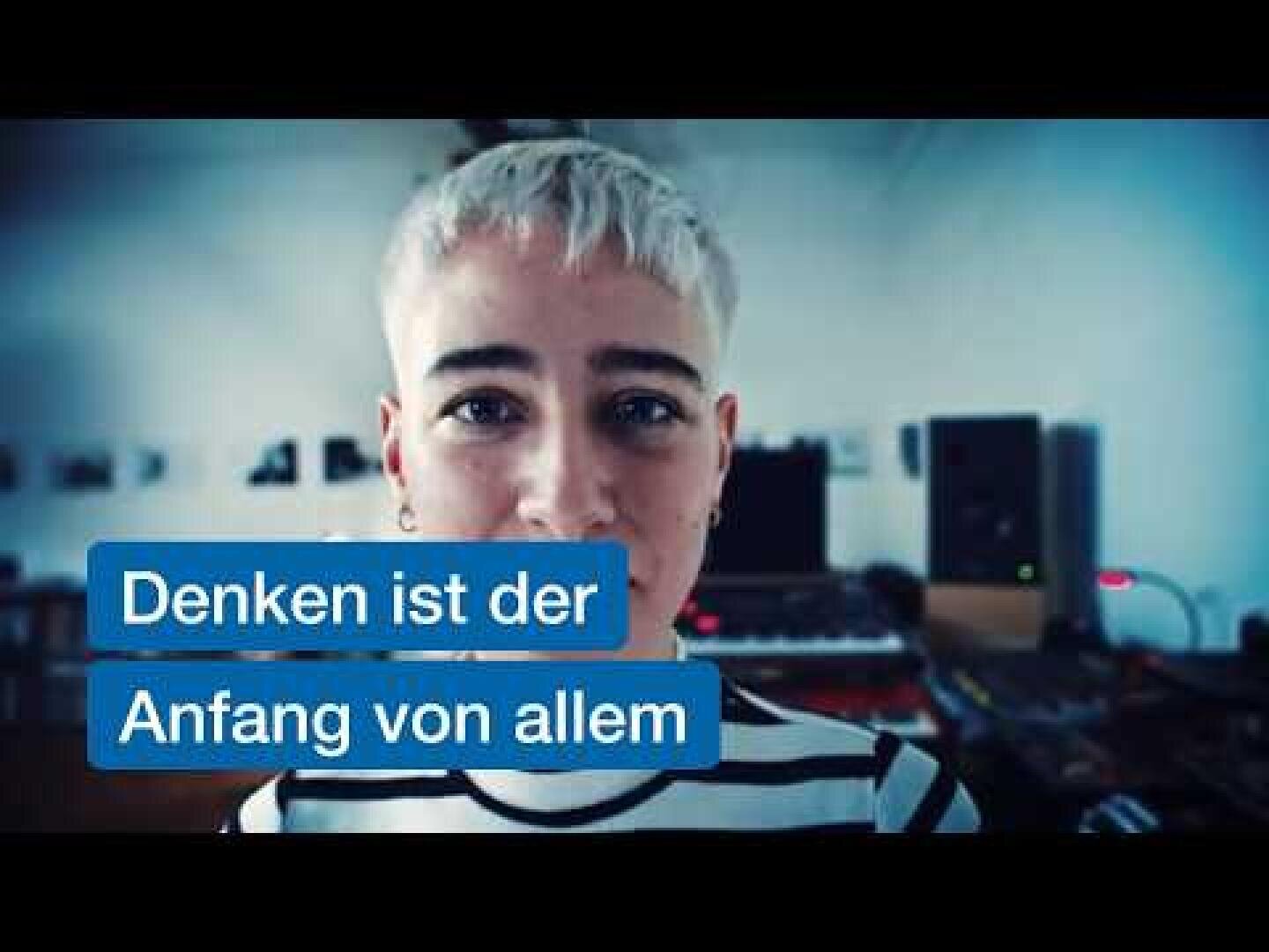 Eine Person mit kurzen platinblonden Haaren und einem gestreiften Hemd steht in einem Raum mit Musikinstrumenten. Auf dem Bild steht auf Deutsch: „Denken ist der Anfang von allem.“.