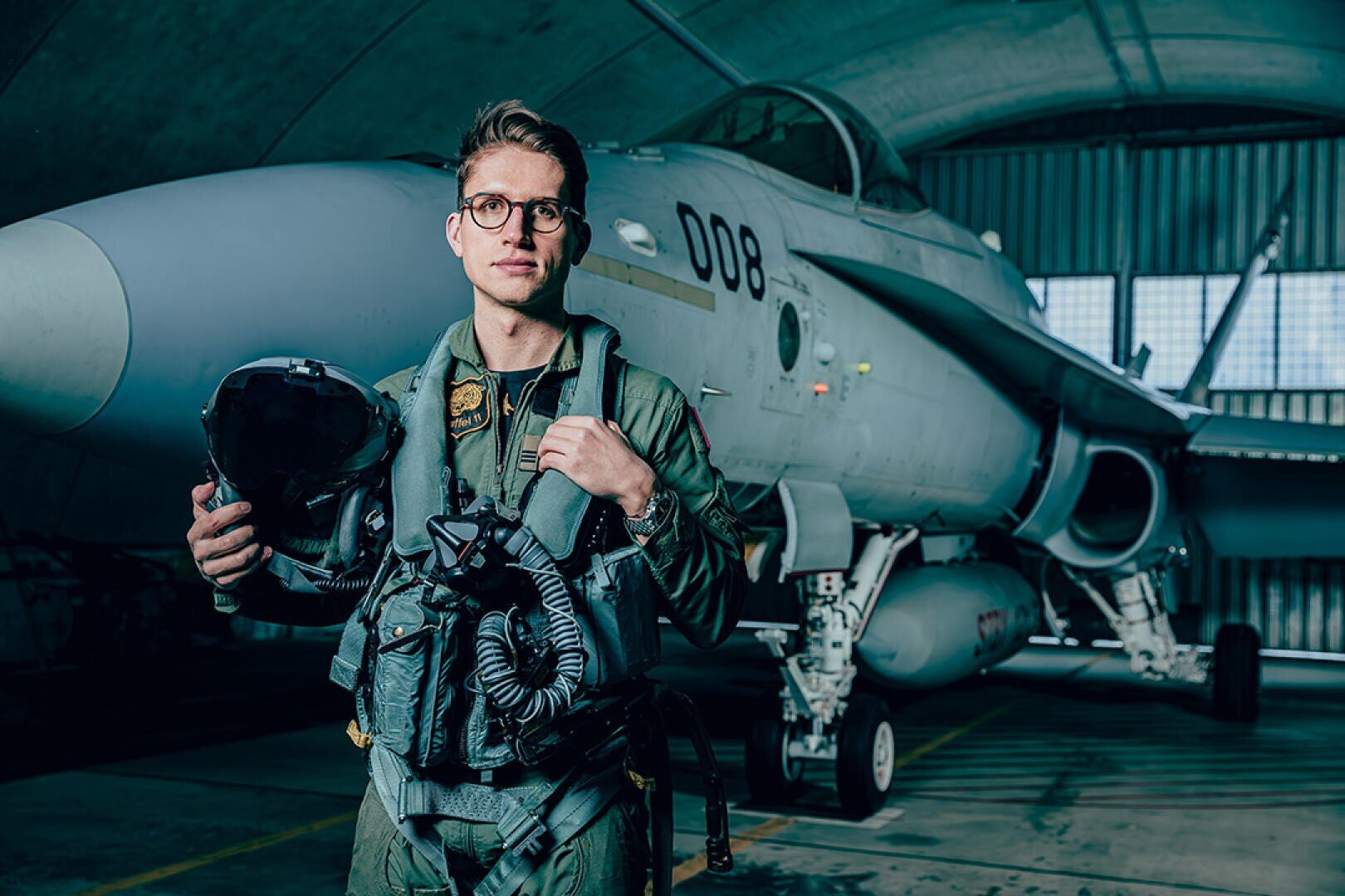 Ein Kampfpilot in einem grünen Fluganzug steht vor einem Jet in einem Hangar, hält einen Helm und blickt zuversichtlich nach vorne. Der Jet hat die Aufschrift 008 auf seiner Nase.