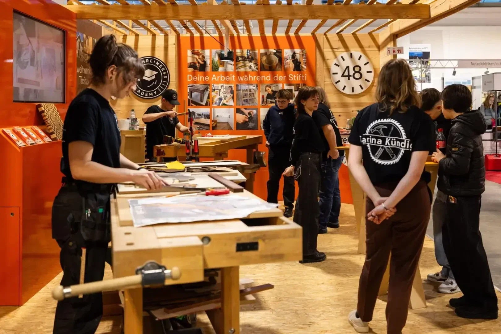 In einer Tischlerei mit Holzbänken, Werkzeugen und Fotos an den Wänden arbeiten und unterhalten sich Menschen. Im Hintergrund sind eine große Uhr und Informationstafeln zu sehen.