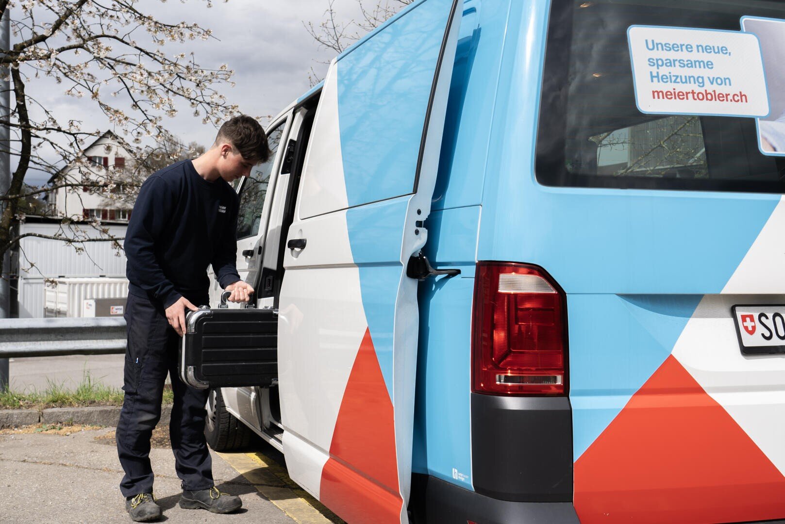 Ein dunkel gekleideter Mann entlädt einen schwarzen Werkzeugkasten aus einem bunten Lieferwagen, der am Straßenrand geparkt ist. Der Lieferwagen ist blau, orange und weiß gestaltet und wirbt mit einem Schild für energieeffizientes Heizen.