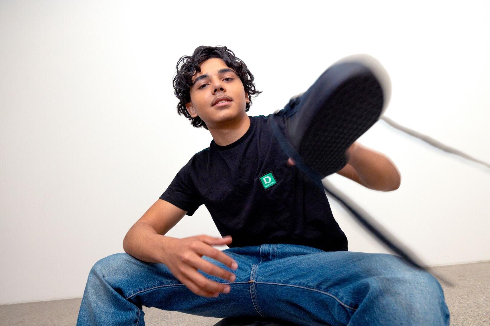 Eine junge Person mit lockigem Haar, die ein schwarzes T-Shirt und blaue Jeans trägt, sitzt da und hebt einen Fuß in Richtung Kamera, als würde sie einen Turnschuh anziehen, mit einem entspannten und lässigen Gesichtsausdruck.