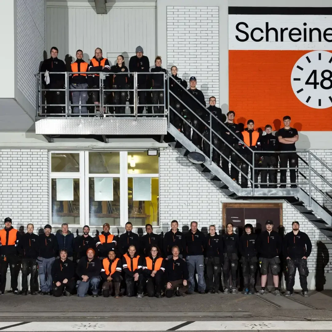 Eine große Gruppe von Menschen in schwarzen und orangefarbenen Arbeitsuniformen posiert auf einer Außentreppe und vor einem weißen Backsteingebäude mit der Aufschrift „Schreiner“ und einer Uhrengrafik, die die Zahl 48 anzeigt.