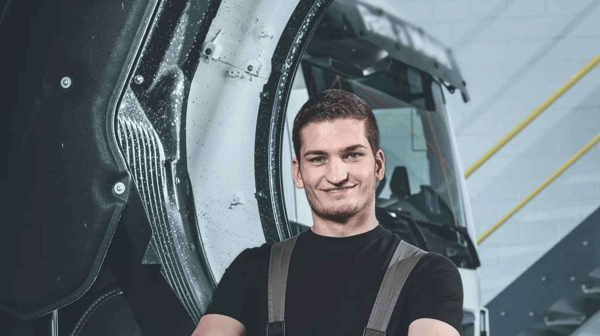 Ein junger Mechaniker in schwarzem Hemd und Latzhose steht selbstbewusst mit verschränkten Armen vor einem großen Lastwagen in einer Werkstatt.