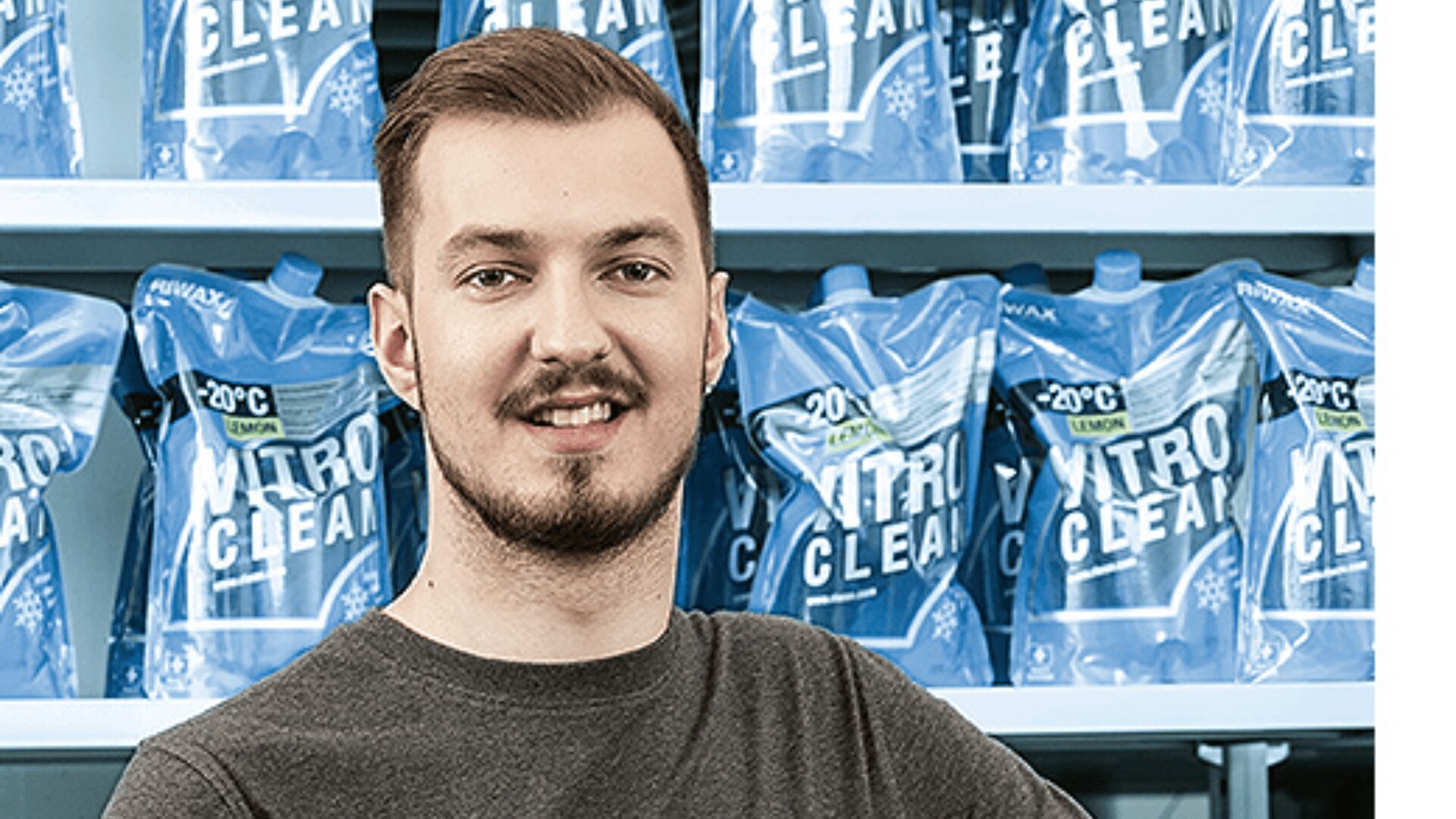 Ein Mann mit kurzen braunen Haaren und gestutztem Bart steht lächelnd vor einem Regal mit blauen Verpackungen mit der Aufschrift NITRO CLEAN.