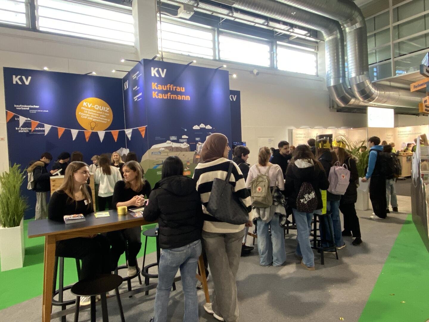 Bei einer Veranstaltung in einer Halle versammelt sich eine Gruppe von Menschen um einen Stand mit der Aufschrift „KV Kaufrau Kaufmann“. Einige sitzen an Tischen und unterhalten sich, während andere in einer Schlange stehen; im Hintergrund sind Informationstafeln und Banner zu sehen.