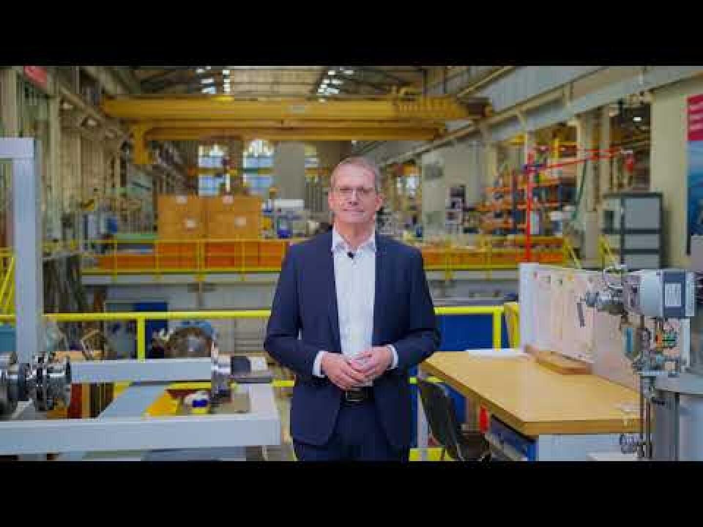 Ein Mann im Anzug steht in einer industriellen Fabrikumgebung, in der Maschinen, Geräte und Regale im Hintergrund zu sehen sind. Er blickt mit gefalteten Händen in einem geräumigen, gut beleuchteten Arbeitsbereich in die Kamera.