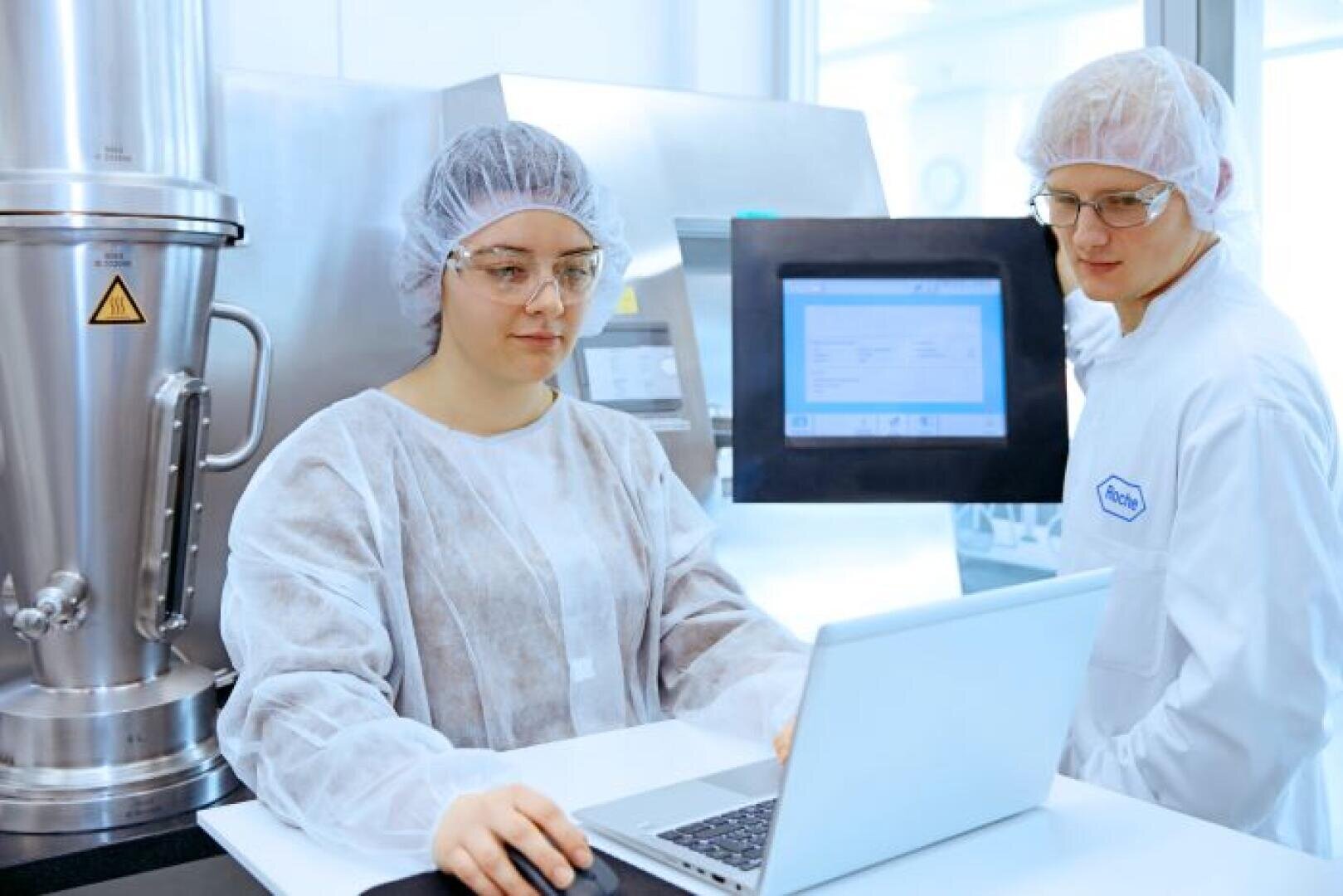 Zwei Wissenschaftler in Laborschutzkleidung und Haarnetzen arbeiten in einer sauberen, modernen Laborumgebung an einem Laptop neben industriellen Geräten und einem Touchscreen-Monitor.