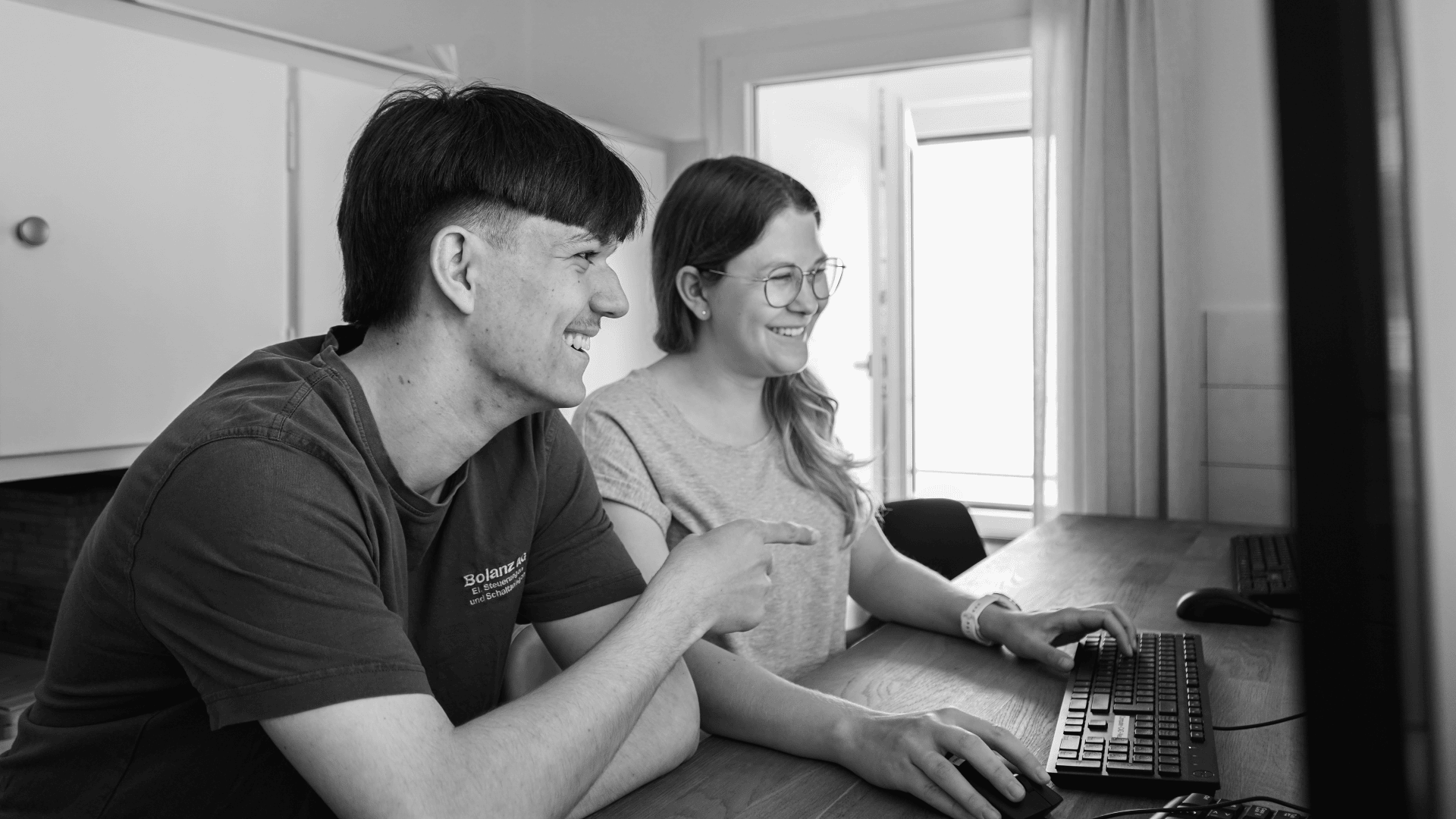 Zwei junge Erwachsene, ein Mann und eine Frau, sitzen nebeneinander an einem Schreibtisch und benutzen einen Computer. Beide lächeln und schauen auf den Monitor in einem hellen Raum mit natürlichem Licht.