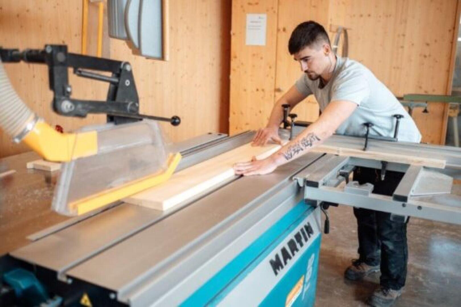 Ein Mann schneidet in einer Werkstatt mit einer Tischsäge ein Holzbrett zu. Er konzentriert sich darauf, das Holz durch die Säge zu führen, die mit einer Schutzvorrichtung versehen ist. Die Werkstatt hat Holzwände und natürliches Licht.