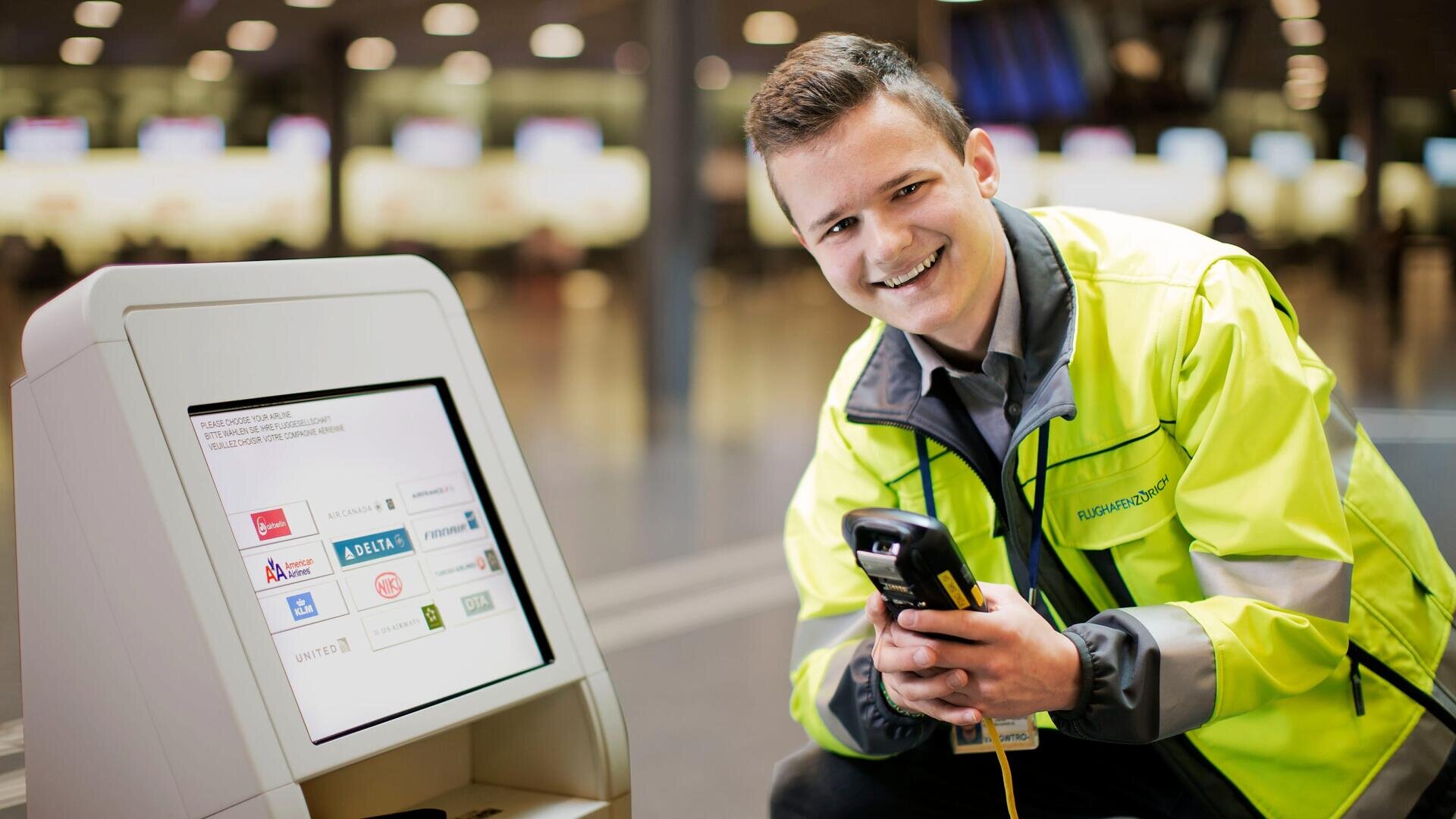 Ein lächelnder Flughafenmitarbeiter in einer gelben Warnweste kniet neben einem Selbstbedienungskiosk und hält einen Handscanner in der Hand. Auf dem Bildschirm des Kiosks sind die Logos der Fluggesellschaften zu sehen, und im Hintergrund ist ein geschäftiges Terminal zu sehen.