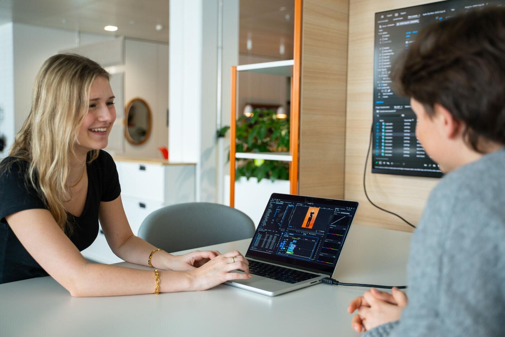 Zwei Personen sitzen an einem Tisch, lächeln und unterhalten sich. Zwischen ihnen ist ein Laptop aufgeklappt, auf dem Daten und Grafiken angezeigt werden. Ein großer Monitor an der Wand zeigt ähnliche Daten an, was auf eine gemeinsame Arbeits- oder Studiensitzung schließen lässt.
