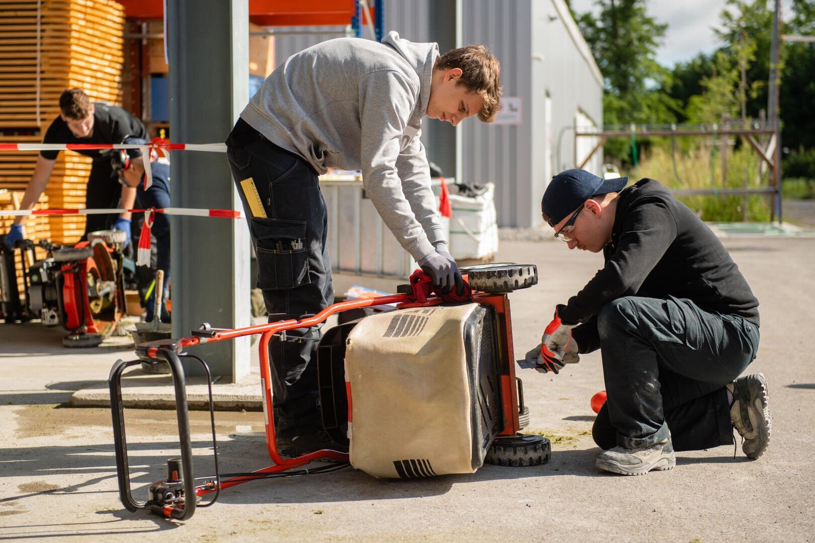 Zwei Männer reparieren einen Rasenmäher im Freien; einer hält den Mäher fest, während der andere mit einem Werkzeug an der Unterseite arbeitet. Um sie herum sind Werkzeuge und Geräte zu sehen.