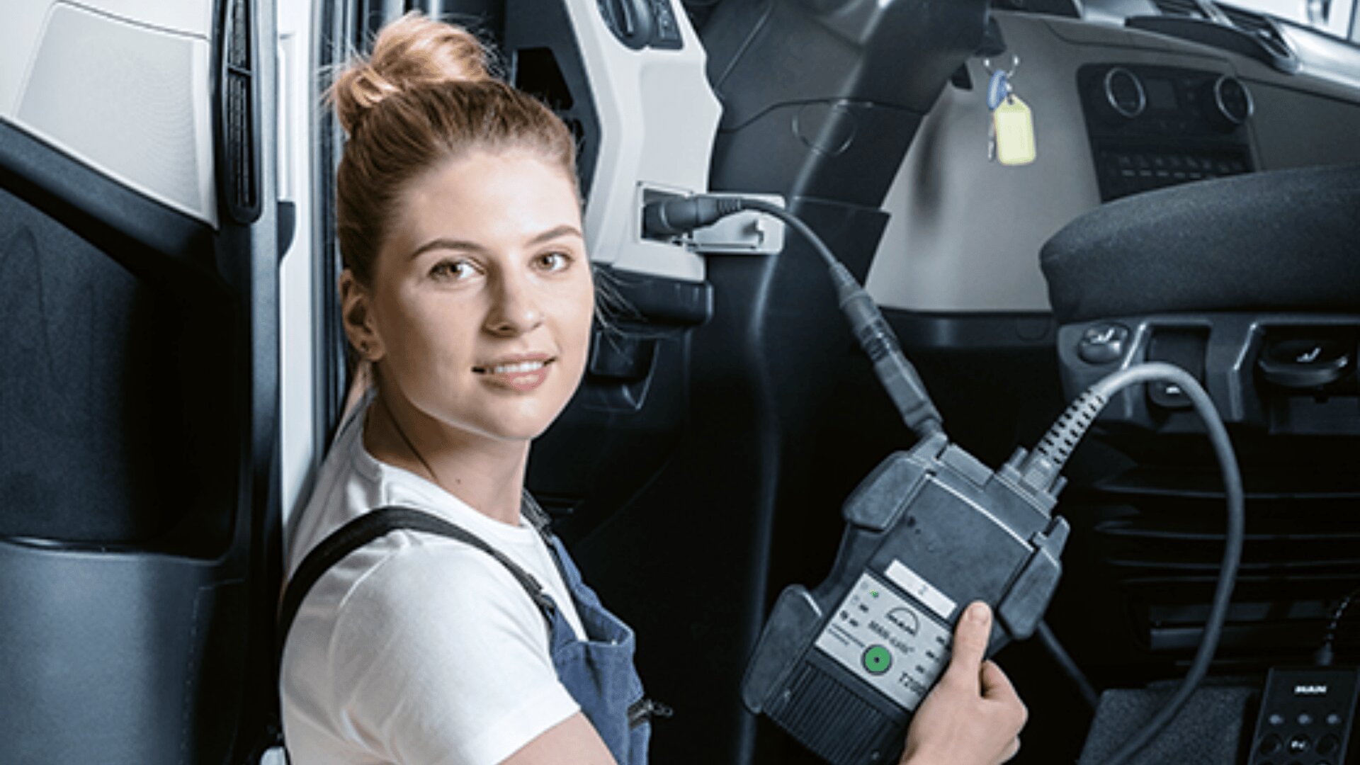 Eine Frau in einem Overall sitzt in einer Autotür und hält ein Kfz-Diagnosegerät in der Hand, das an das Armaturenbrett des Fahrzeugs angeschlossen ist, schaut in die Kamera und lächelt.
