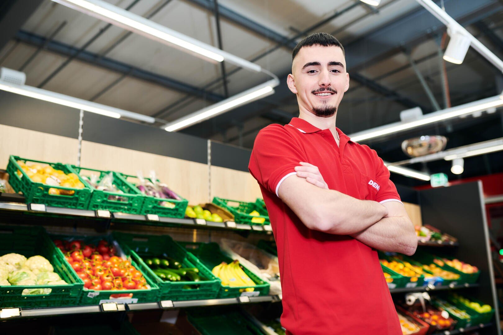 Ein junger Mann in einem roten Polohemd steht mit verschränkten Armen und einem Lächeln vor den Auslagen für Frischwaren in einem Lebensmittelladen. Gemüse wie Paprika, Gurken und Zucchini sind in den Regalen hinter ihm zu sehen.