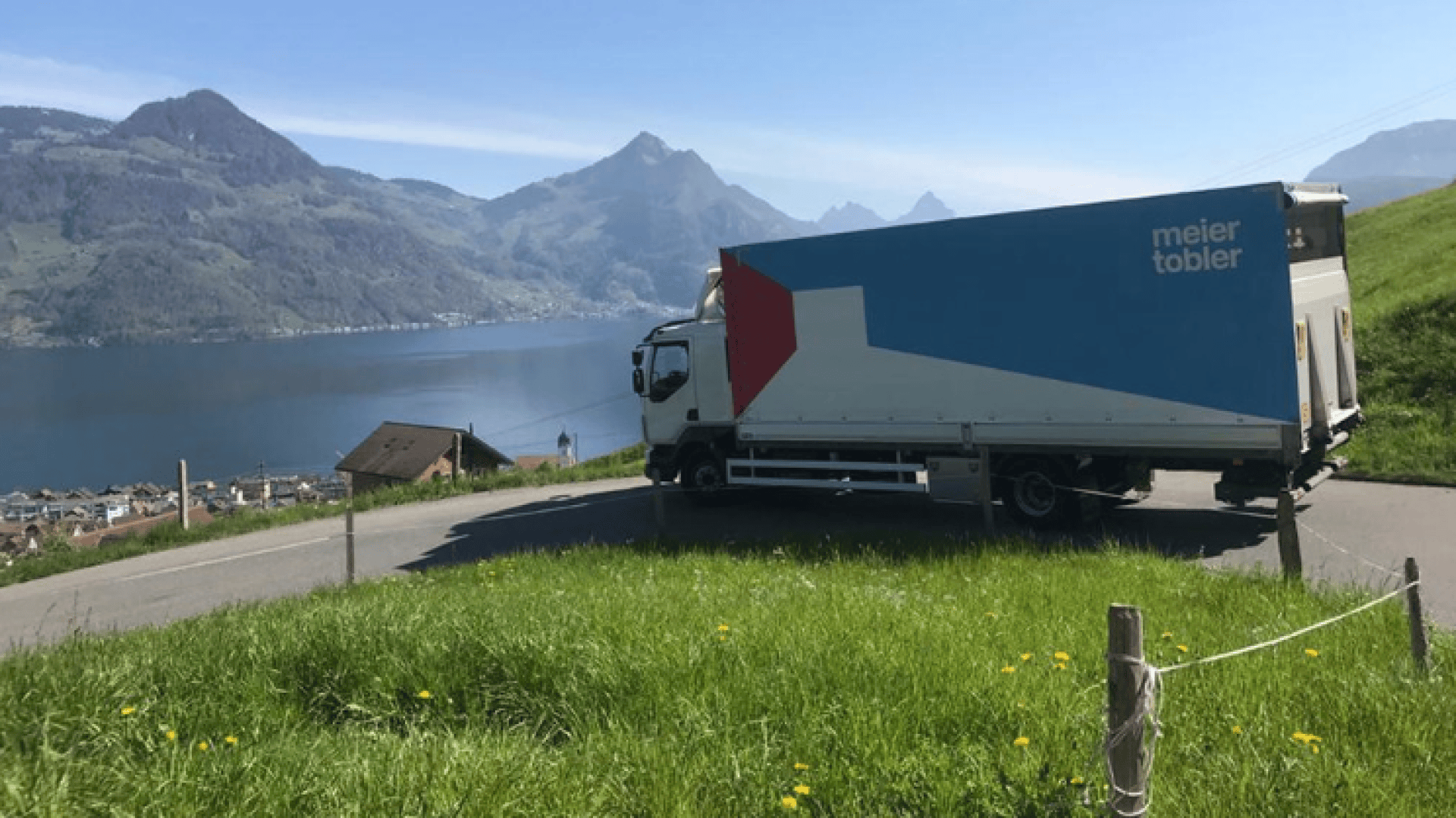 Ein großer Lieferwagen mit dem Schriftzug meier tobler steht auf einer kurvenreichen Straße mit Blick auf einen See, Berge und ein kleines Dorf, mit grünem Gras und Wildblumen im Vordergrund.