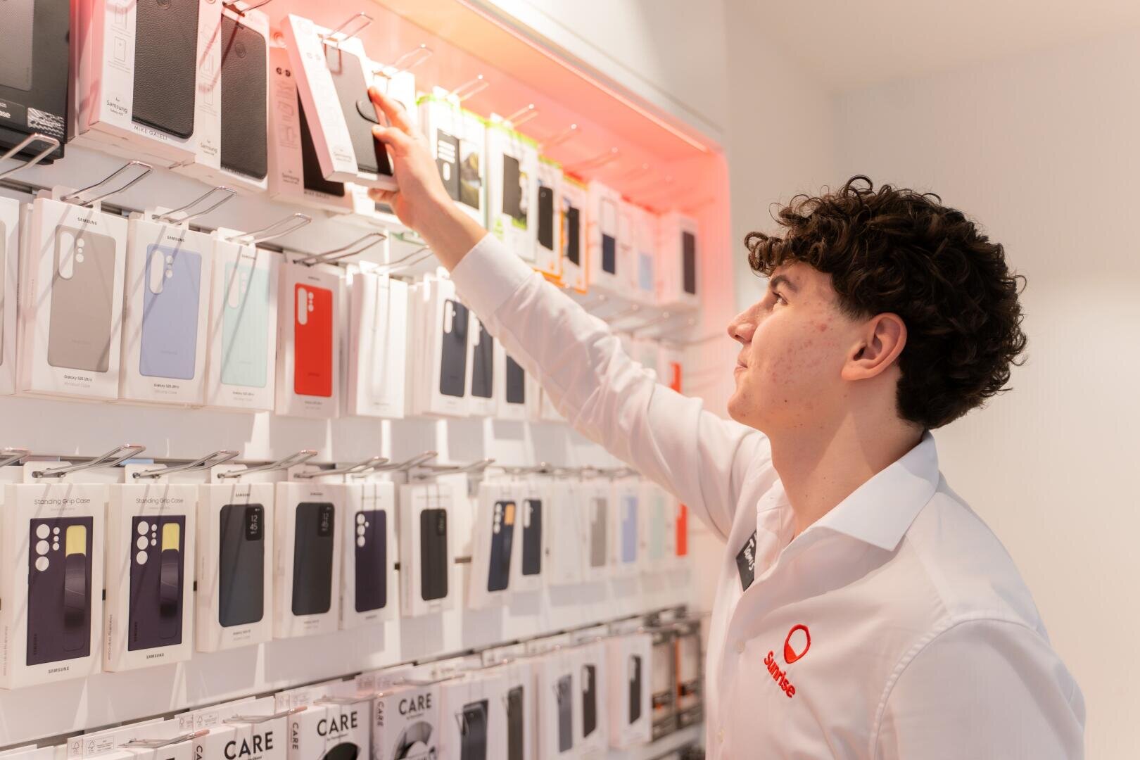 Eine Person in einem weißen Hemd greift in einem Einzelhandelsgeschäft nach einer Smartphonehülle aus einer Ausstellungswand mit verschiedenen Handyhüllen.