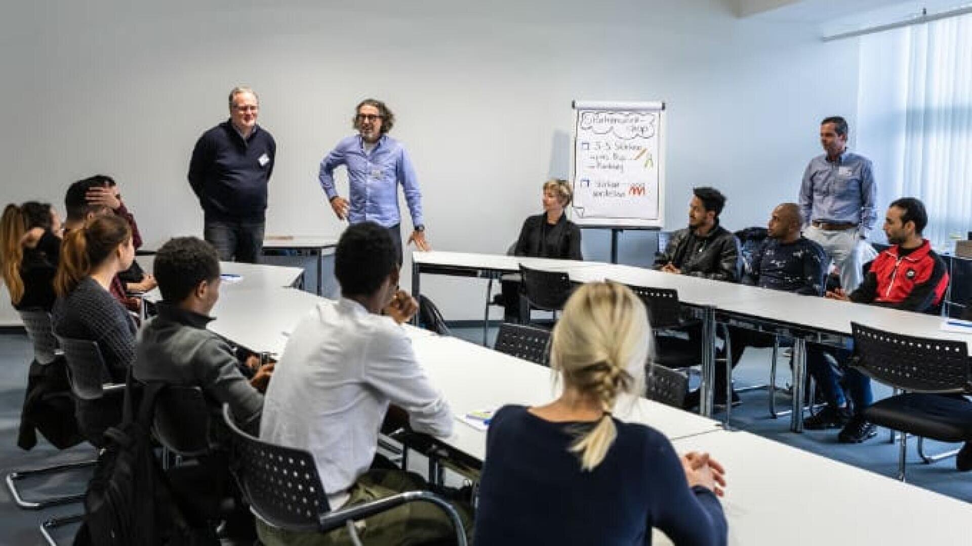 Eine Gruppe von Menschen sitzt in einem Besprechungsraum um einen U-förmigen Tisch und hört drei stehenden Männern zu. Im Hintergrund ist ein Flipchart mit Notizen zu sehen. Die Atmosphäre wirkt kollaborativ.