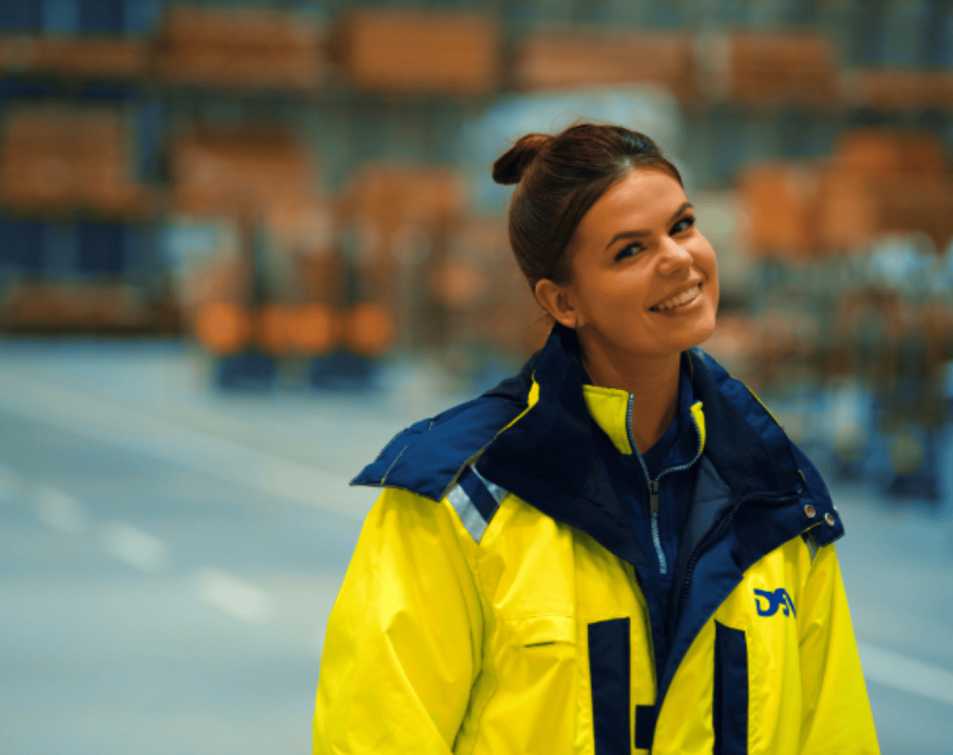 Eine Frau in einer leuchtend gelben Warnweste steht lächelnd in einem Lagerhaus, im Hintergrund sind Regale und Industrieanlagen verschwommen zu sehen.