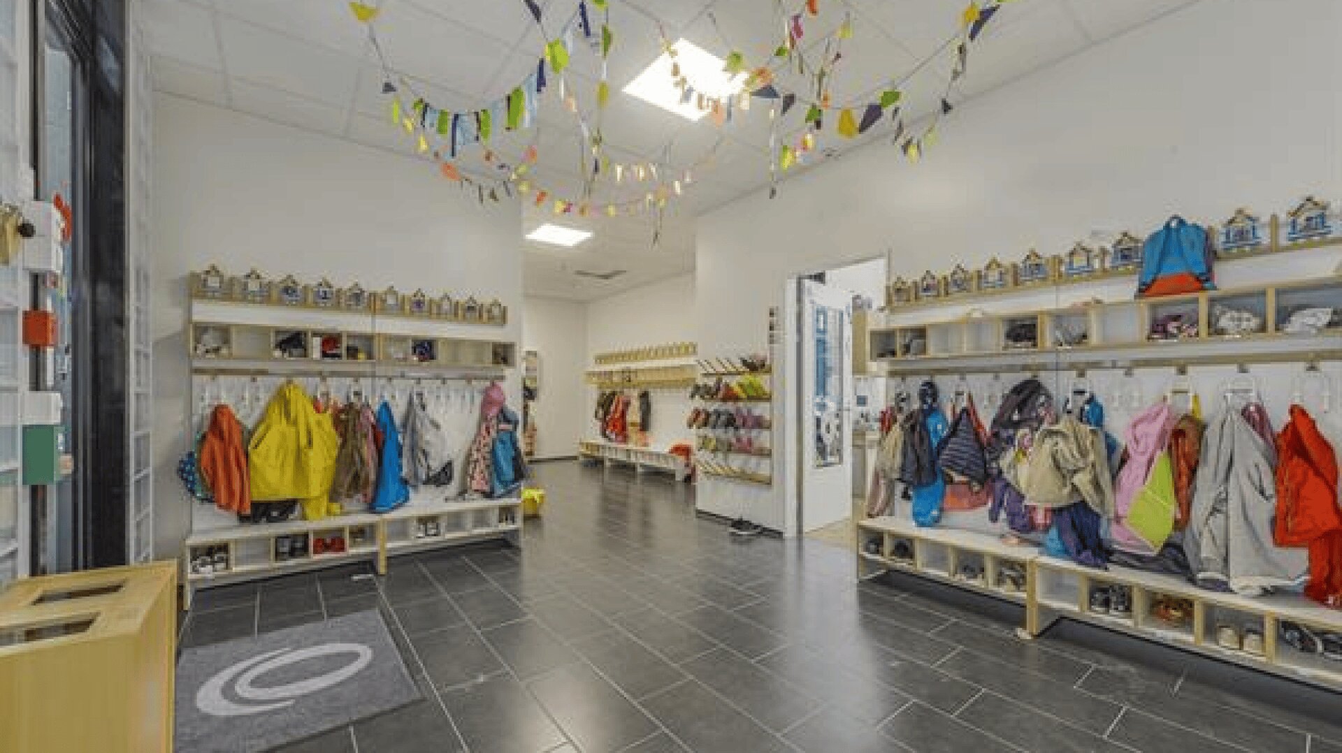 Eine helle, geordnete Garderobe mit Reihen von Fächern und Haken für bunte Kinderjacken und -schuhe. Von der Decke hängen Wimpel, und durch die großen Fenster auf der linken Seite fällt Tageslicht ein.