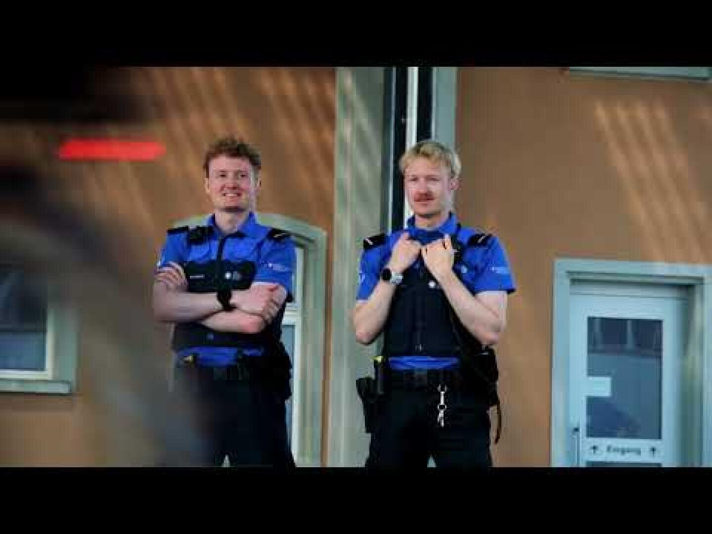 Zwei uniformierte Polizisten stehen vor einem Gebäude mit beigefarbenen Wänden. Sie lächeln und wirken entspannt. Beide tragen blaue Hemden und schwarze Westen; einer hat die Arme verschränkt, der andere hält die Träger seiner Weste fest. Im Hintergrund sind Türen zu sehen.