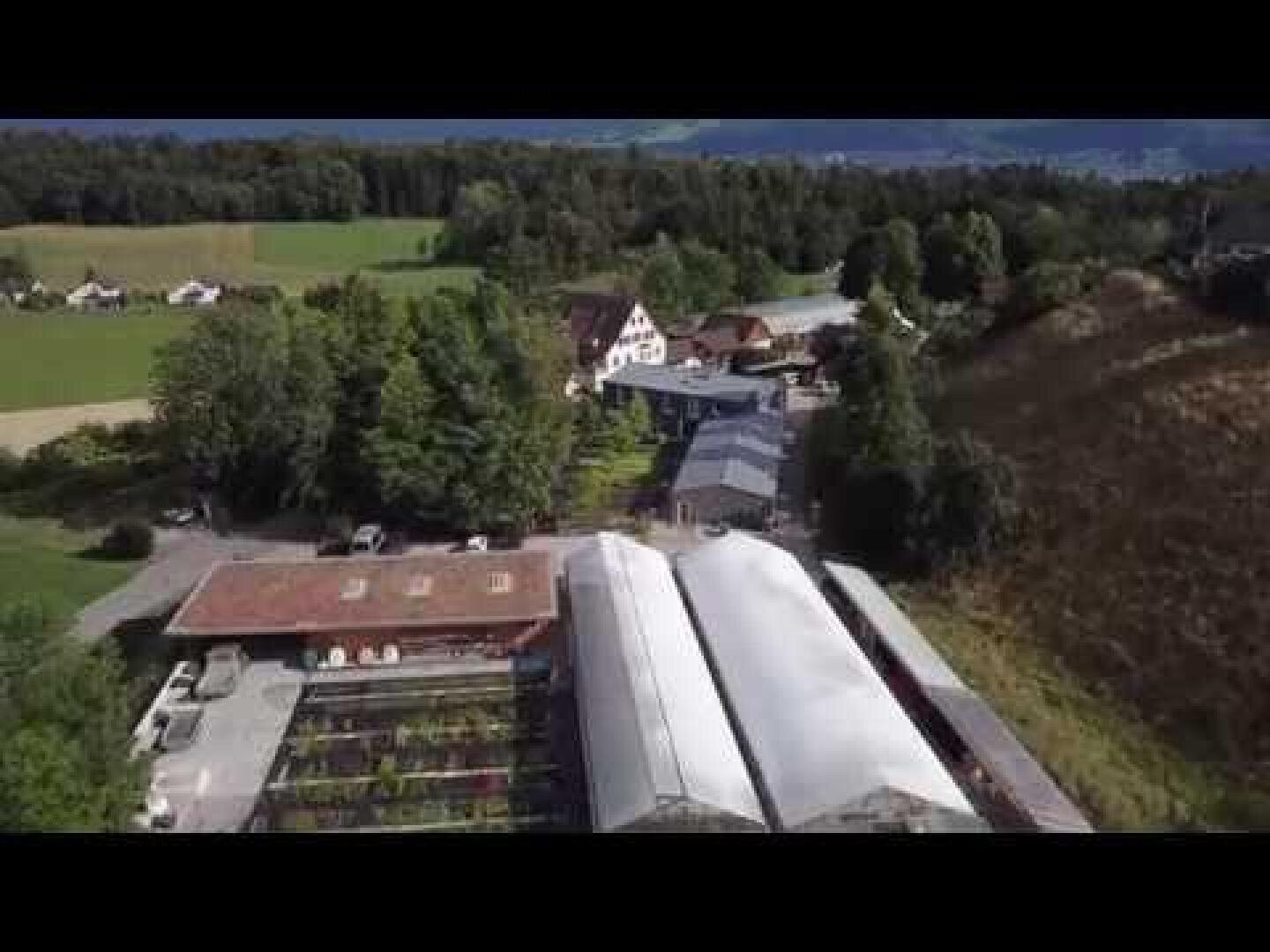 Luftaufnahme einer ländlichen Gegend mit Gewächshäusern, Bäumen und mehreren Gebäuden, umgeben von Feldern und bewaldeten Hügeln im Hintergrund.