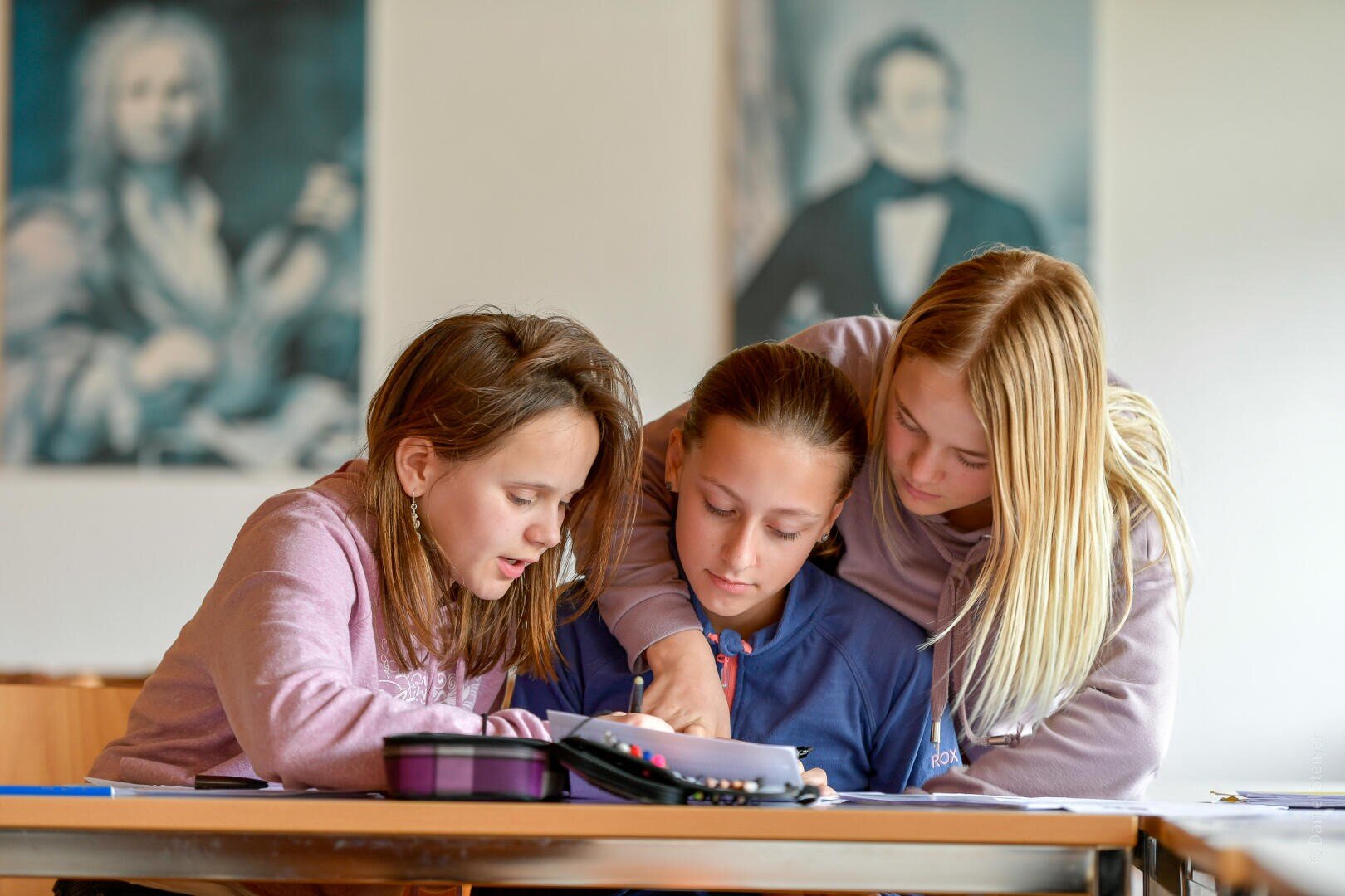 Drei Mädchen sitzen zusammen an einem Tisch, arbeiten an Schulaufgaben und teilen sich ein Heft. Zwei der Mädchen lehnen sich eng aneinander, während das dritte Mädchen schreibt. An der Wand im Hintergrund hängen unscharfe Porträts.