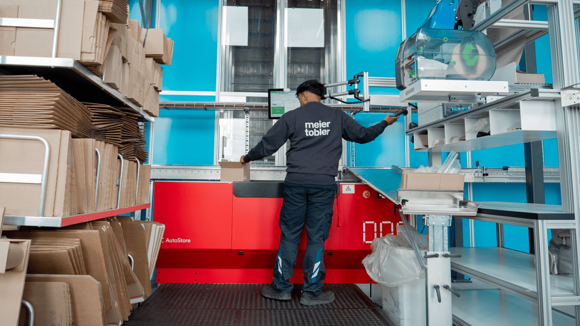 Eine Person mit einer Jacke von meier tobler steht vor einem roten Arbeitsplatz und bearbeitet Pakete in einem modernen, organisierten Lager mit Regalen aus Kartons und blauen Wänden.