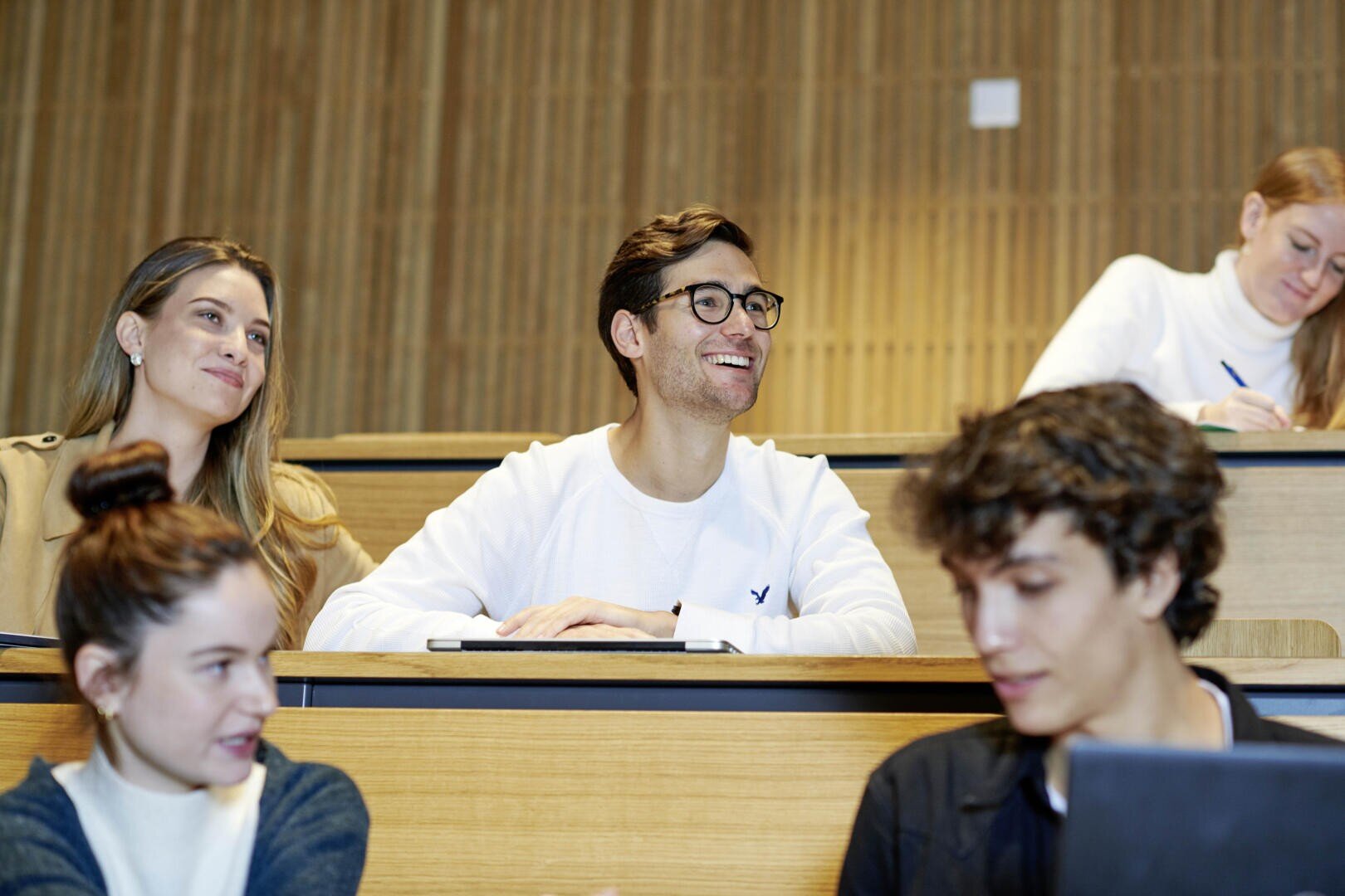 Studenten sitzen in einem Vorlesungssaal, einige lächeln und machen sich Gedanken, andere machen sich Notizen. Die Gruppe wirkt aufmerksam und konzentriert, im Hintergrund ist eine Holzverkleidung zu sehen.