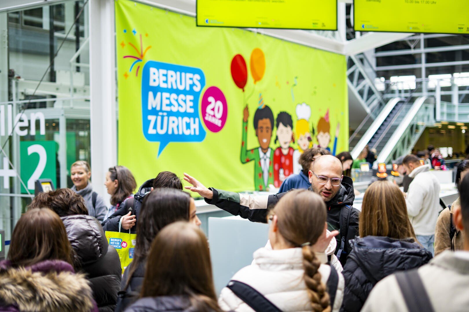Bei einer Indoor-Veranstaltung mit einem bunten Banner mit der Aufschrift Berufs Messe Zürich herrscht reges Treiben. Die Menschen sind in Gespräche vertieft, und die Atmosphäre wirkt lebendig und energiegeladen.