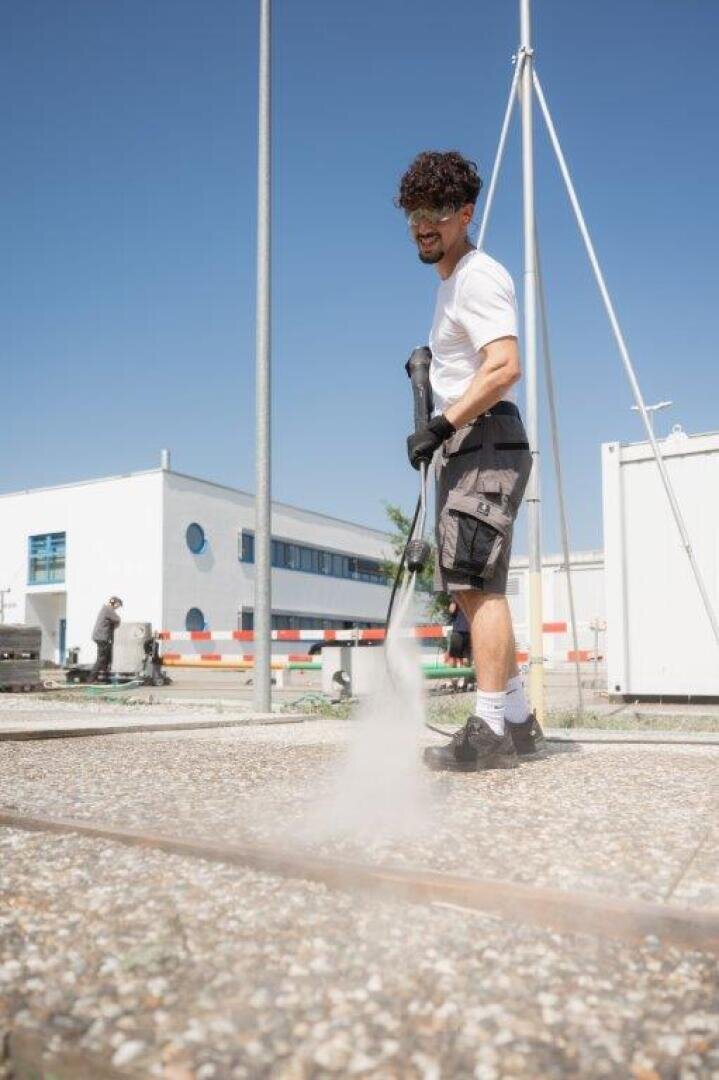Ein Mann in weißem Hemd, Shorts, Handschuhen und Sicherheitsschuhen reinigt an einem sonnigen Tag mit einem Hochdruckreiniger eine Fläche im Freien, mit Industriegebäuden und einem klaren blauen Himmel im Hintergrund.
